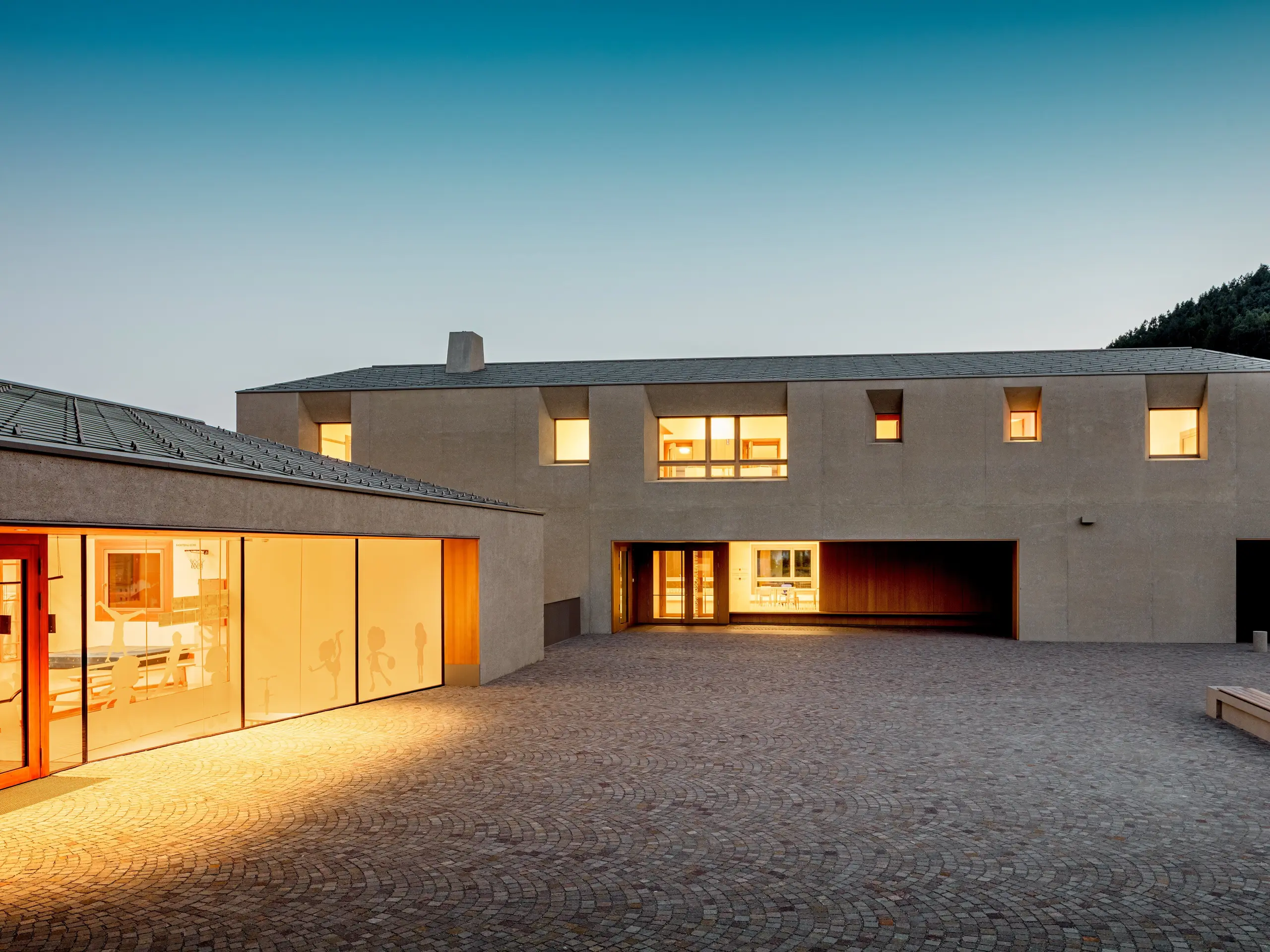 Au crépuscule, le centre éducatif de Riffian diffuse une lueur chaleureuse sur la cour pavée. Les losanges de toiture PREFA 44 × 44, en finition P.10 Bronze, coiffent le jardin d’enfants et l’école primaire d’un revêtement en aluminium d’une grande précision ; ils allient une architecture épurée à une durabilité à toute épreuve, tout en apportant une touche élégante et sereine au pied des versants montagneux du Tyrol du Sud.