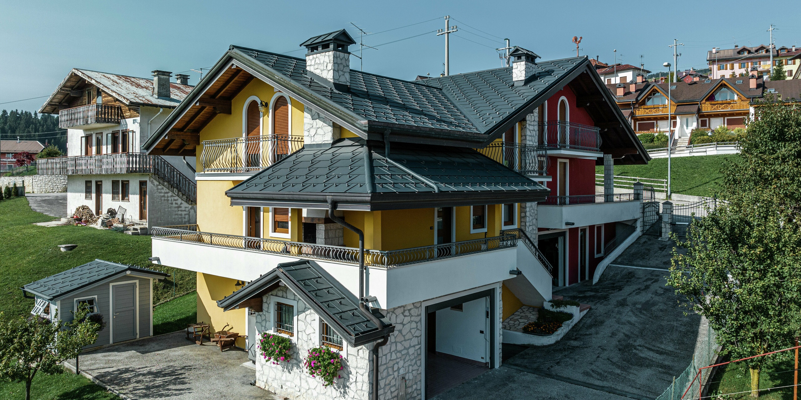 Maison individuelle à Gallio de style architectural alpin avec toiture moderne en R.16 PREFA en P.10 gris sombre. Les panneaux de toiture PREFA garantissent une solution de toiture durable, résistante aux intempéries et visuellement attrayante. La combinaison harmonieuse de la façade jaune-rouge, des éléments en pierre naturelle et des accents en bois confère à la maison un caractère accueillant. Le système de gouttière PREFA en P.10 gris sombre assure un drainage fiable de l'eau. Un excellent exemple d’architecture esthétique et fonctionnelle dans un environnement alpin.
