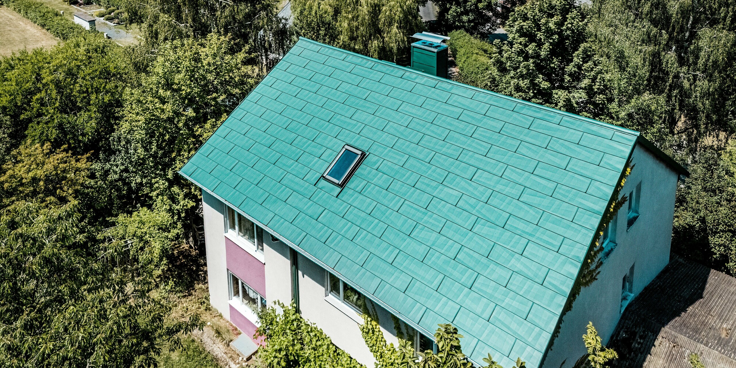 Maison individuelle modernisée à Schwarzenbach-Martinlamitz, dotée d'une toiture en panneaux PREFA FX.12 vert mousse. La photographie aérienne révèle la toiture en aluminium, installée avec précision, à la texture distinctive et agrémentée d'un puits de lumière intégré, nichée dans un écrin de verdure. L'association saisissante du vert mousse de la toiture et du blanc violacé de la façade confère à la maison une allure unique. La toiture PREFA séduit par sa durabilité, sa résistance aux intempéries et son confort sans entretien – idéale pour des rénovations élégantes et haut de gamme.