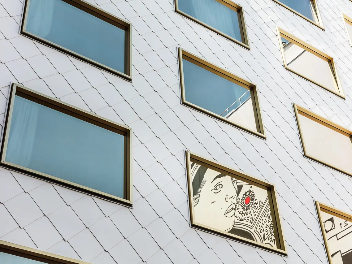 Détail de la façade de l'hôtel "THE ROCK Radisson RED Vienna", habillée de losanges de toit et de murs PREFA 44 × 44 en P.10 blanc pur. Parmi les nombreuses fenêtres à vitrage miroir et aux cadres dorés, une fenêtre se distingue par une œuvre d'art encastrée, représentant le portrait d'une personne avec un accent rouge prononcé