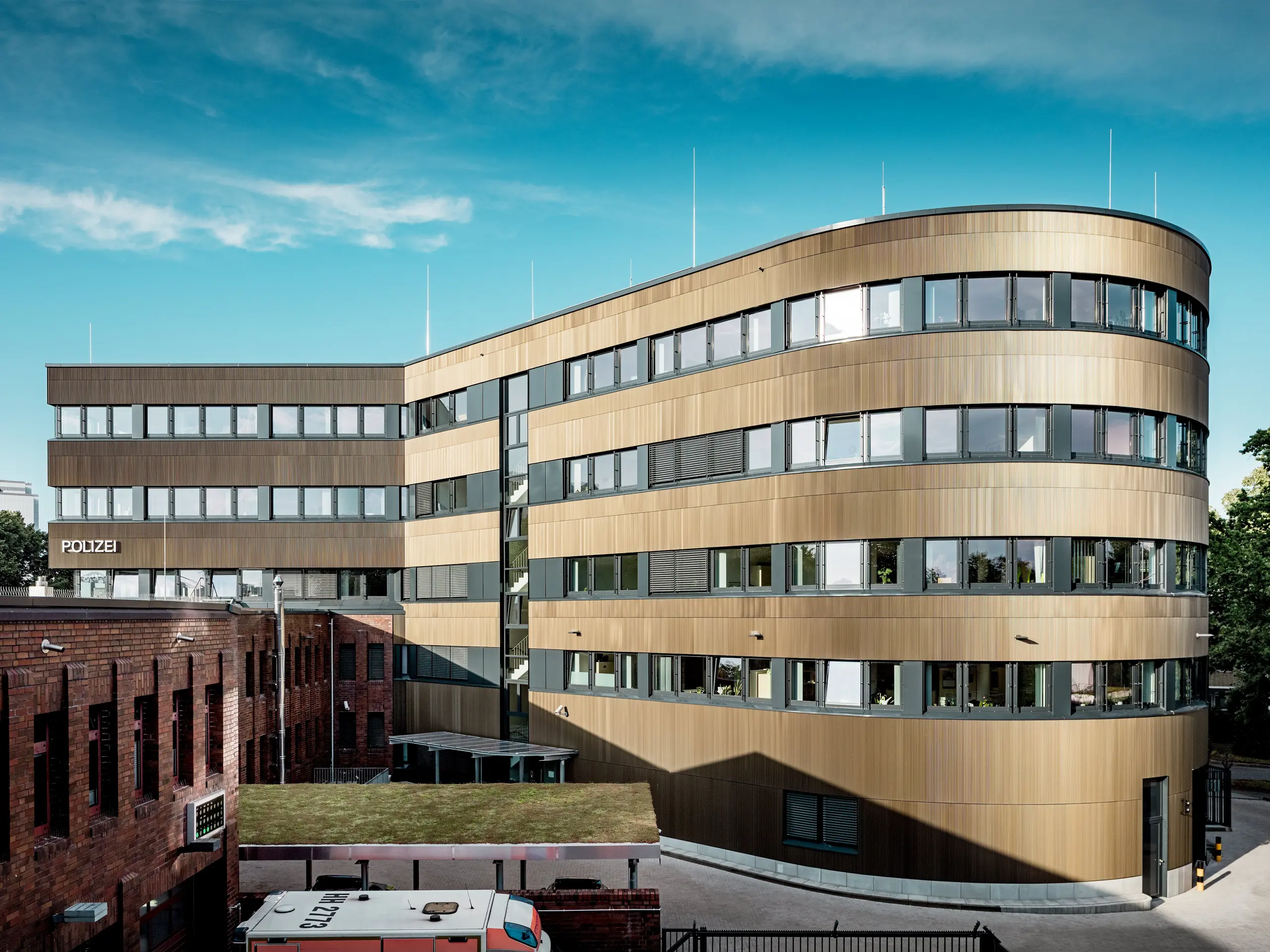 Vue d'ensemble saisissante du commissariat 43 à Hambourg : le nouveau bâtiment incurvé, avec sa façade en aluminium PREFA d'environ 1 100 m² composée de profils extrudés « triangle » couleur bronze moyen, se distingue nettement des bâtiments en briques existants. La surface finement texturée crée de magnifiques jeux d'ombre et de lumière, faisant de ce bâtiment un véritable chef-d'œuvre architectural. Grâce à sa haute résistance aux intempéries, sa protection contre la corrosion, sa durabilité et ses exigences minimales d'entretien, les solutions en aluminium PREFA incarnent une architecture de façade durable, fonctionnelle et esthétique.