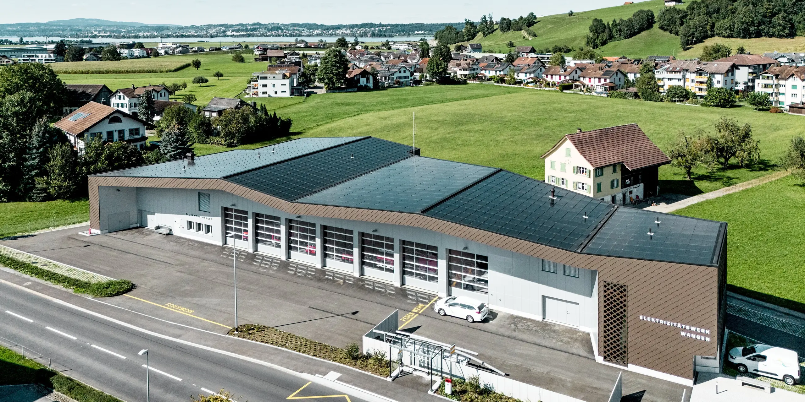Vue aérienne du bâtiment abritant la caserne des pompiers et la centrale électrique de Wangen, en Suisse, niché dans un paysage vallonné pittoresque. Le revêtement de façade remarquable, composé de losanges PREFA 29 x 29 en coloris P.10 Brun, crée des effets architecturaux saisissants et s'harmonise avec les panneaux composites en aluminium clair P.10 Prefa Blanc. Ces produits en aluminium durables se distinguent par leur haute résistance aux intempéries, leur faible besoin d'entretien et leur capacité à constituer une enveloppe de bâtiment moderne et durable.