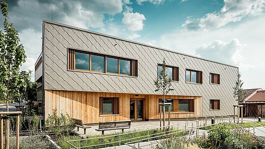 La façade de la crèche d'Alzenau révèle une entrée principale accueillante, harmonieusement composée de losanges de façade PREFA couleur bronze (44 x 44 cm) et d'une façade verticale en bois au rez-de-chaussée. Le revêtement en aluminium, à la structure épurée, dessine un élégant motif d'échelle, rythmé par de larges fenêtres à encadrements en bois et persiennes verticales. Au premier plan, des allées accessibles, des parterres de fleurs et des bancs soulignent la qualité de l'espace. L'alliance de l'aluminium résistant, du bois naturel et d'un aménagement paysager soigné fait de ce bâtiment un exemple architectural de construction durable, fonctionnelle et esthétique pour les établissements scolaires utilisant les systèmes de façade PREFA.