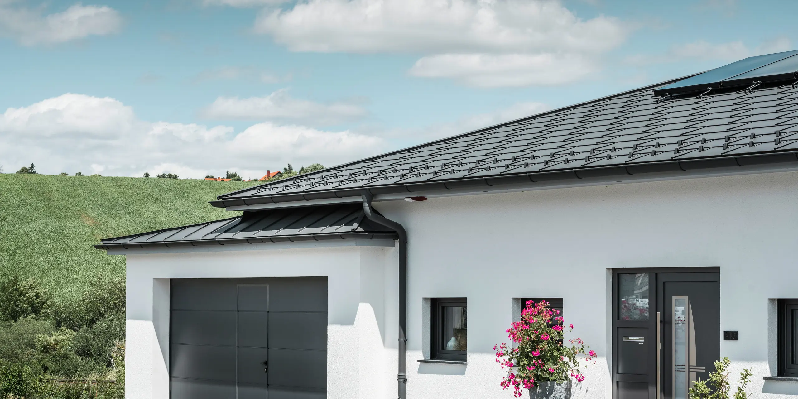 La photo montre la face endroit d'une maison individuelle à Ilmenau, en Thuringe, avec une toiture PREFA élégante en P.10 gris sombre. La tuile R.16 confère à la maison un aspect moderne, tandis que le système de drainage de toiture PREFA intégré, avec sa gouttière demi-ronde et son tuyau de descente gris sombre, garantit une évacuation efficace de l'eau de pluie. Le garage, équipé de PREFALZ gris sombre, s'intègre harmonieusement dans l'esthétique générale et souligne la qualité et le style des produits en aluminium PREFA.
