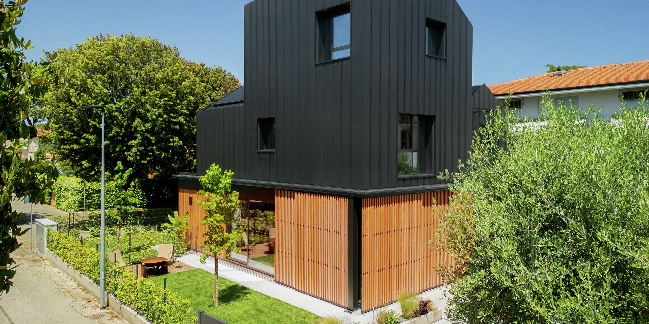 Vue de rue d'une maison jumelée moderne à Cattolica : une élégante combinaison de lattes de bois au rez-de-chaussée et d'une façade en aluminium PREFALZ gris noir. Les lignes épurées et la surface de haute qualité PREFA offrent durabilité, protection contre la corrosion et une architecture intemporelle sous un ciel ensoleillé.