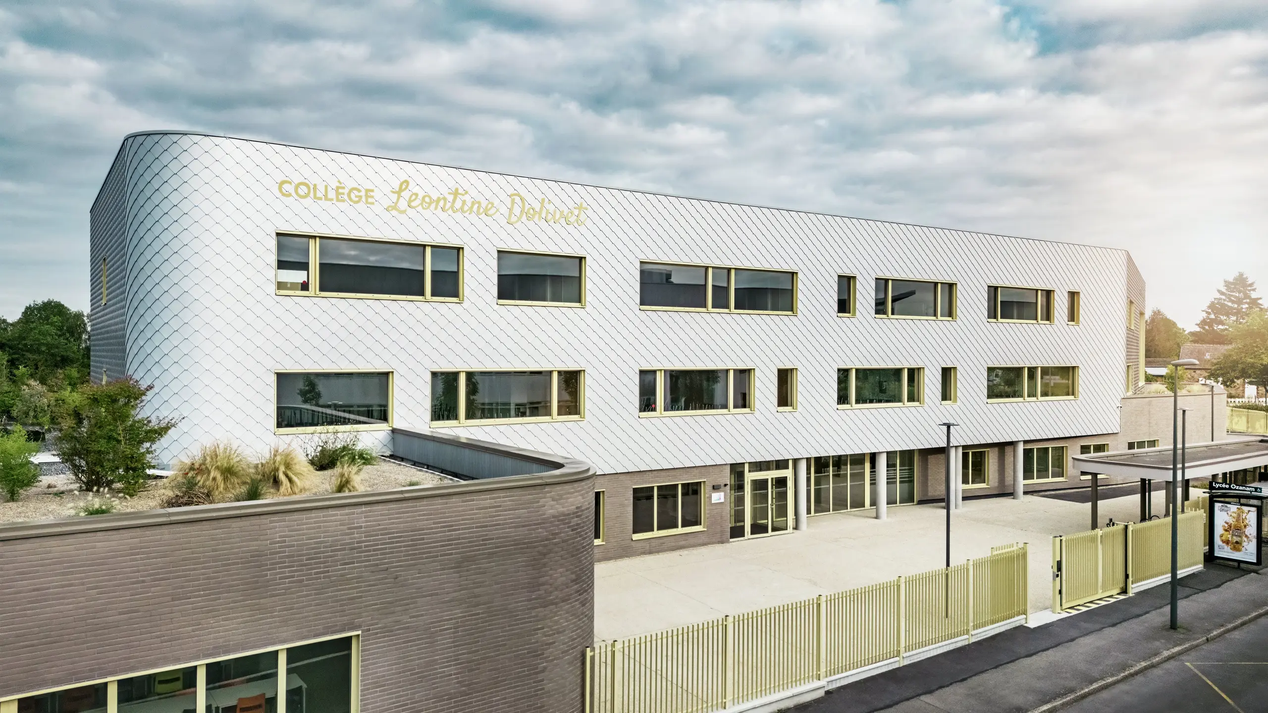 Vue de face du Collège Léontine Dolivet à Cesson-Sévigné, dont la façade moderne en aluminium est composée de losanges PREFA argent métallisé. La surface irisée confère au bâtiment une impression de légèreté, tandis que les lignes épurées et les larges fenêtres assurent une transparence optimale. Des encadrements de fenêtres jaunes apportent des touches de dynamisme. Au premier plan, on aperçoit une place lumineuse, une clôture et la rue adjacente. La solution de façade PREFA, durable et résistante, allie esthétique, résistance aux intempéries et durabilité – un véritable atout architectural, témoignant d'une élégance raffinée.