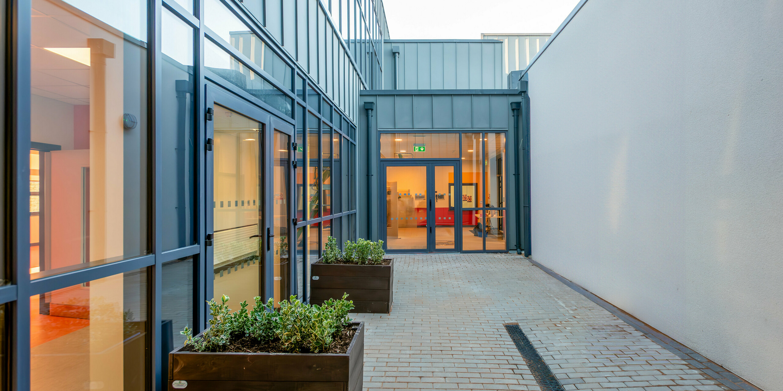 Passage moderne de Scoil Mhuire Naofa à Carrigtwohill, Irlande. De grandes façades vitrées et une façade en aluminium PREFALZ en P.10 gris souris assurent une architecture contemporaine et baignée de lumière. Des jardinières et des matériaux légers créent une atmosphère accueillante.