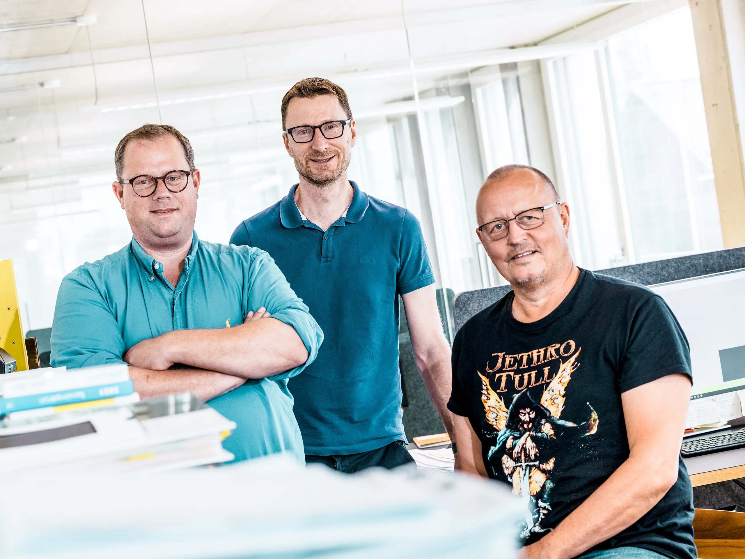 Photo de l'équipe de Kaundbe Architects dans leurs bureaux modernes : les trois architectes posent avec assurance devant leurs postes de travail et leurs documents techniques. Au premier plan, des piles de plans et de dossiers de projets ; à l'arrière-plan, des écrans d'ordinateur affichent des plans de construction. La scène reflète la compétence, la collaboration et la précision technique – des valeurs essentielles dans la conception et la réalisation de solutions de façade avec les produits en aluminium PREFA.