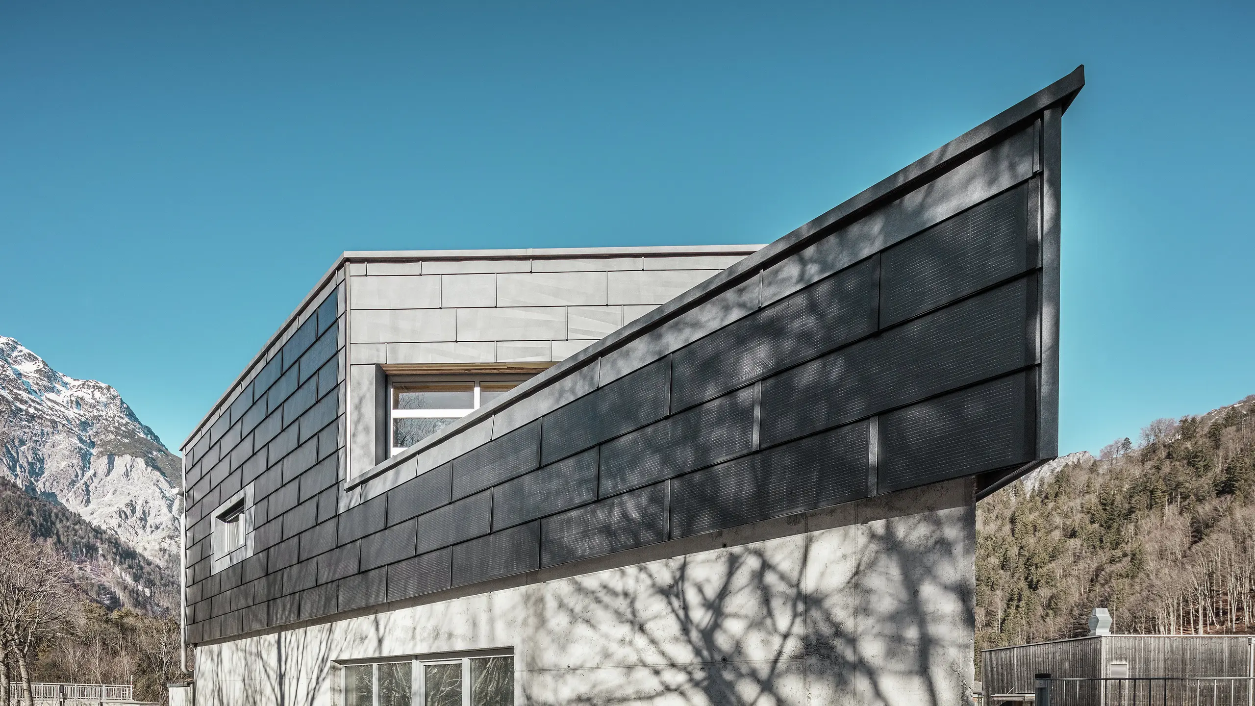 Le bâtiment de l'entreprise métallurgique RÜMA, dans le Vorarlberg, impressionne par sa façade moderne en tuiles solaires PREFA et béton apparent. Entouré de montagnes et d'arbres, dont les ombres se projettent sur le mur, il se dresse sous un ciel bleu et dégagé. L'angle pointu du bâtiment et les panneaux de façade PREFA FX.12 soulignent encore son allure imposante.