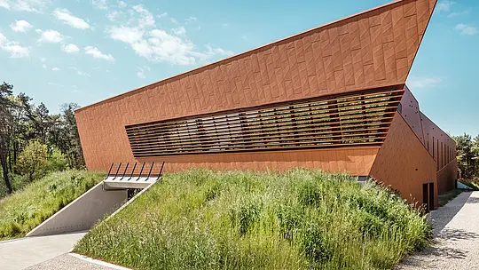 La remarquable station de pompage d'eau potable WML de Lottum se distingue par son toit incliné et sa façade en aluminium PREFA Siding.X aux formes dynamiques et à l'aspect chaleureux et rouillé. La surface facettée crée un jeu d'ombre et de lumière saisissant, tandis que les persiennes horizontales ajoutent de la profondeur architecturale. Niché dans un talus naturel, le bâtiment se dresse comme un repère sculptural. Les panneaux d'aluminium PREFA, durables, légers et recyclables, offrent une résistance exceptionnelle aux intempéries et une stabilité dimensionnelle remarquable, ainsi qu'une enveloppe de bâtiment durable et nécessitant peu d'entretien.