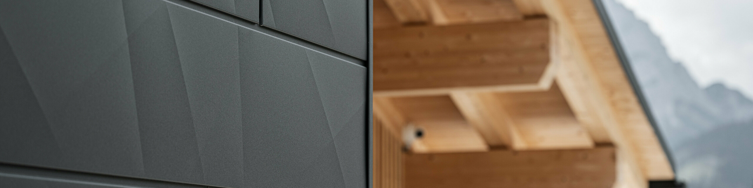 Gros plan sur les panneaux de façade Siding.X en P.10 noir de PREFA sur la Casa Hoffe à Sappada, en Italie. Le système de façade en aluminium est habilement intégré dans un design moderne et abstrait qui contraste agréablement avec les éléments en bois apparents de la charpente de toit. Cette solution de façade innovante s'intègre parfaitement dans l'environnement alpin et allie fonctionnalité, durabilité et esthétique architecturale.