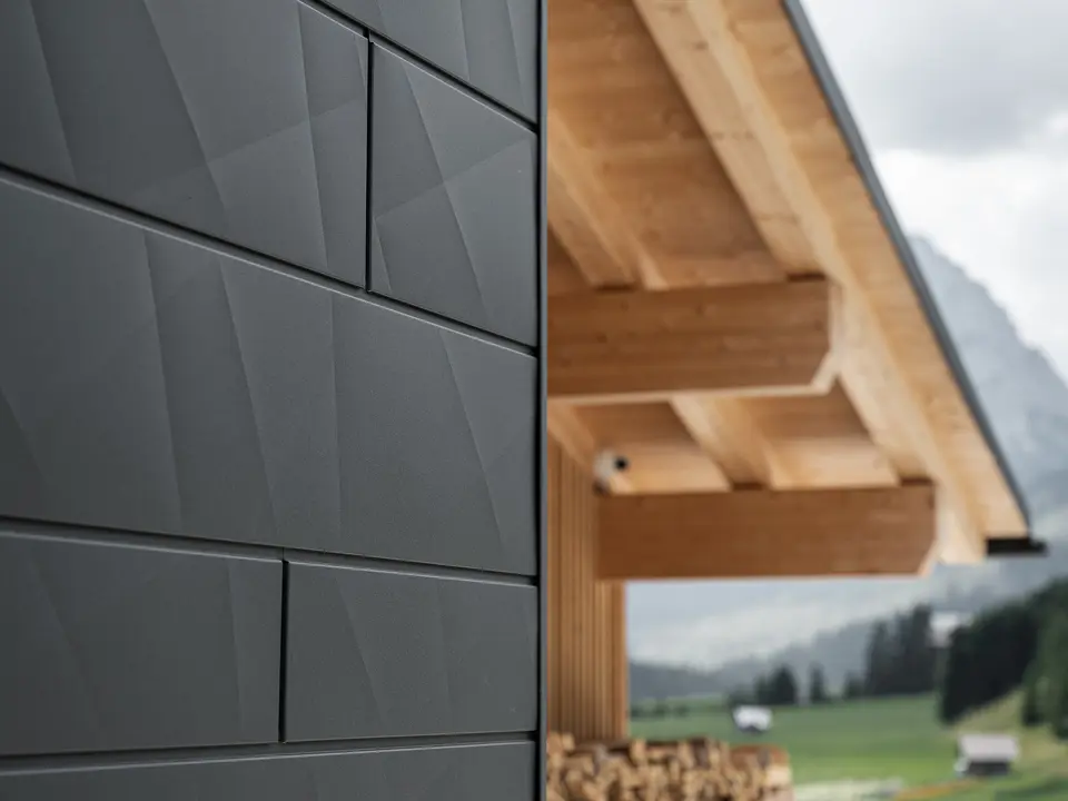 Gros plan sur les panneaux de façade Siding.X en P.10 noir de PREFA à la Casa Hoffe à Sappada, Italie. Le système de façade en aluminium au design moderne et abstrait, crée un contraste intéressant avec les éléments apparents de la charpente en bois. Cette solution de façade innovante, qui s'intègre harmonieusement à l'environnement alpin, combine fonctionnalité, durabilité et culture architecturale esthétique.