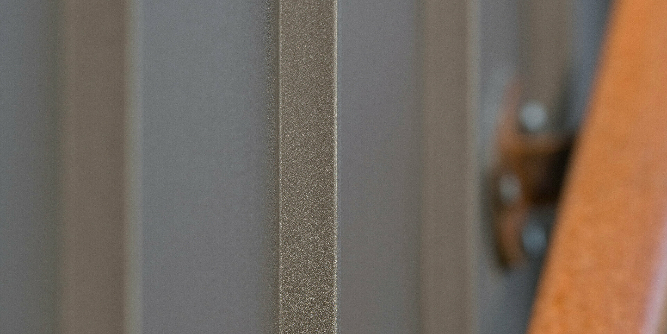 Gros plan d'une façade brun à joints debout posée en bacs verticaux. Le matériau provient de PREFA Aluminium Products GmbH. La texture visible de l'aluminium transmet qualité et robustesse. L'accent mis sur le matériau montre en détail sa texture et ses nuances de couleurs. Une partie floue d'une main courante métallique de couleur rouille complète l'image de droite, créant un contraste par la couleur et la texture.
