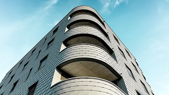Vue dynamique du dessous d'un immeuble résidentiel et commercial moderne, caractérisé par un angle arrondi saisissant et des balcons aux lignes fluides. La façade, aux allures d'écailles, réalisée en bardage PREFA P.10 Gris souris, enveloppe le bâtiment tel une peau d'aluminium finement travaillée, soulignant la douceur de ses courbes. L'homogénéité de la surface met en valeur la pureté des lignes, tandis que la légèreté et la résistance aux intempéries de l'aluminium PREFA garantissent une stabilité dimensionnelle maximale, une grande durabilité et un entretien minimal, même pour les courbes les plus prononcées – un véritable chef-d'œuvre architectural se détachant sur un ciel d'un bleu éclatant.