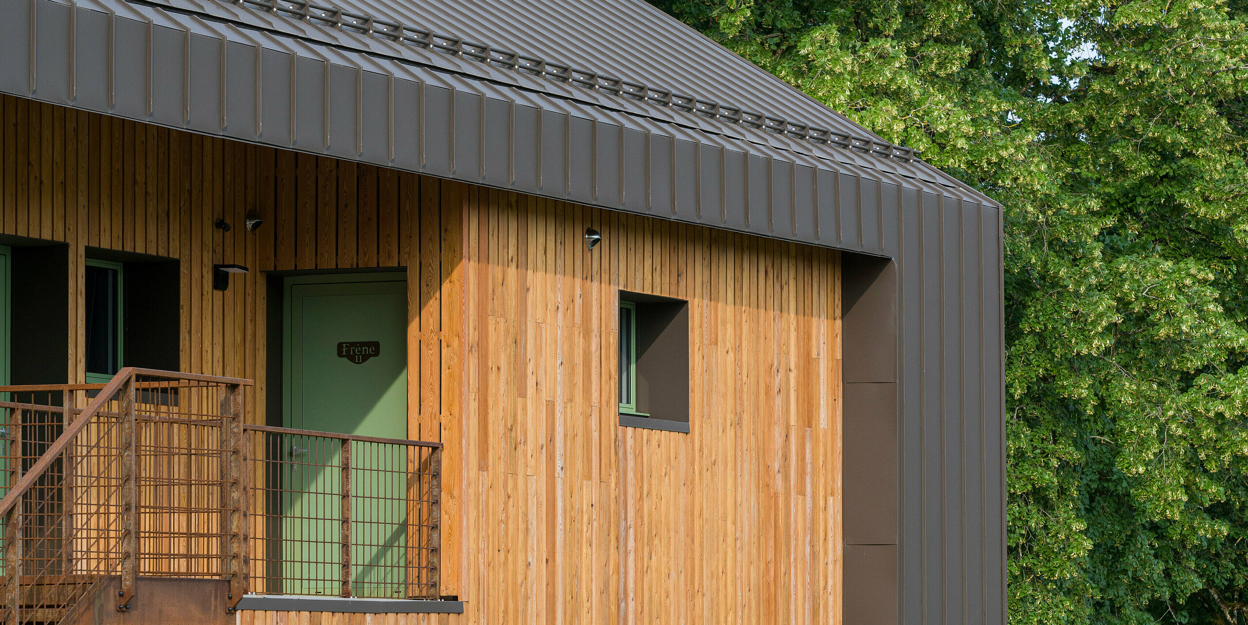 Vue latérale au format portrait de l'Hôtel en Tilleul à Pont du Navoy (France). Le bâtiment présente une remarquable façade en bois associée à des éléments en aluminium constitués de PREFALZ de couleur P.10 brun. Le toit a été entièrement recouvert d'un revêtement à joints debout qui s'étend sur l'avant-toit et décore également les bords de la façade. Il y a quatre chaises et une table en bois devant le bâtiment. L'étage supérieur est bordé par une balustrade métallique. L'arrière-plan est constitué de grands arbres.