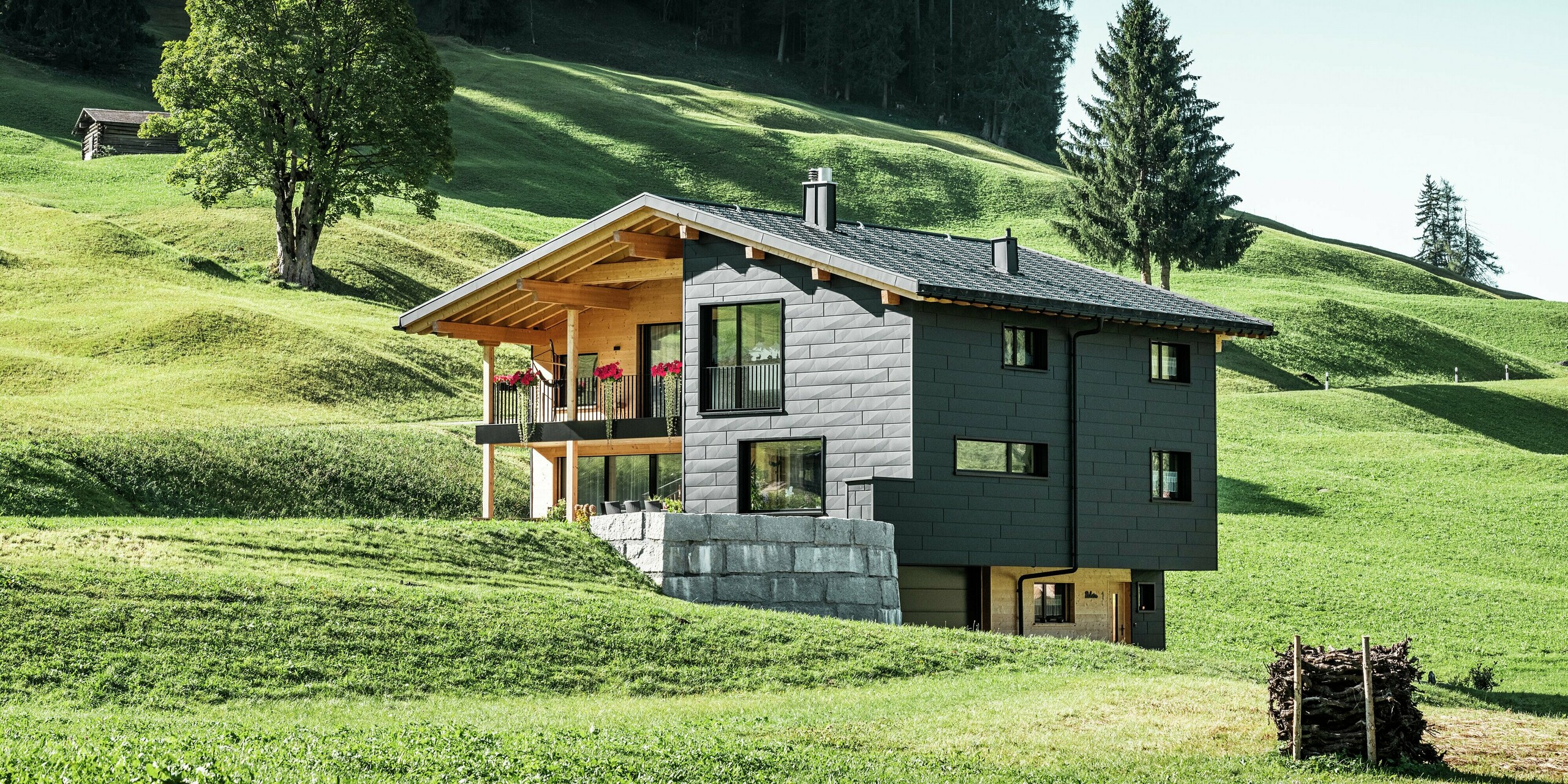 Vue lointaine de la terrasse et du balcon de la maison, conçus en bois et complétant parfaitement la façade robuste en aluminium en PREFA Siding.X en P.10 noir. La façade confère au bâtiment une esthétique moderne et une résistance optimale aux intempéries, tandis que les grandes fenêtres laissent entrer beaucoup de lumière du jour à l'intérieur et encadrent la nature environnante.