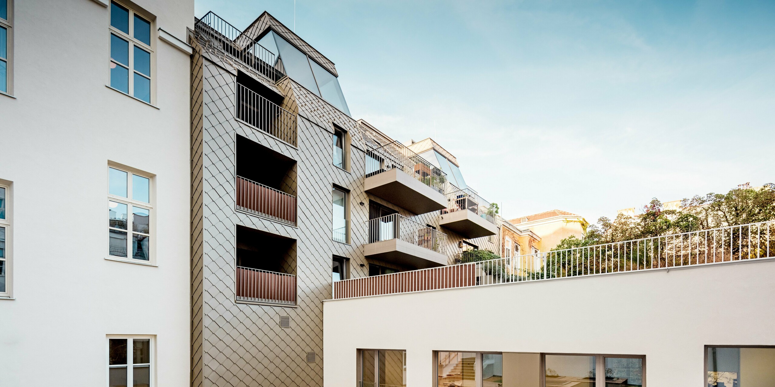 Das Bild zeigt die harmonische Integration der modernen PREFA Rauten 29×29 in Bronze an der Fassade eines Wohngebäudes in Wien. Die stilvollen Rauten verlaufen über die Außenwand und verbinden die modernen Balkone mit der Architektur des Gebäudes. Die Fassade vereint klassische Elemente mit zeitgemäßen Akzenten und passt sich perfekt der Umgebung an. Im Vordergrund ist der Innenhof mit einem minimalistischen Design aus Holzterrassen und Pflanzbeeten zu sehen, was das moderne und durchdachte Gesamtkonzept der Wohnanlage unterstreicht.