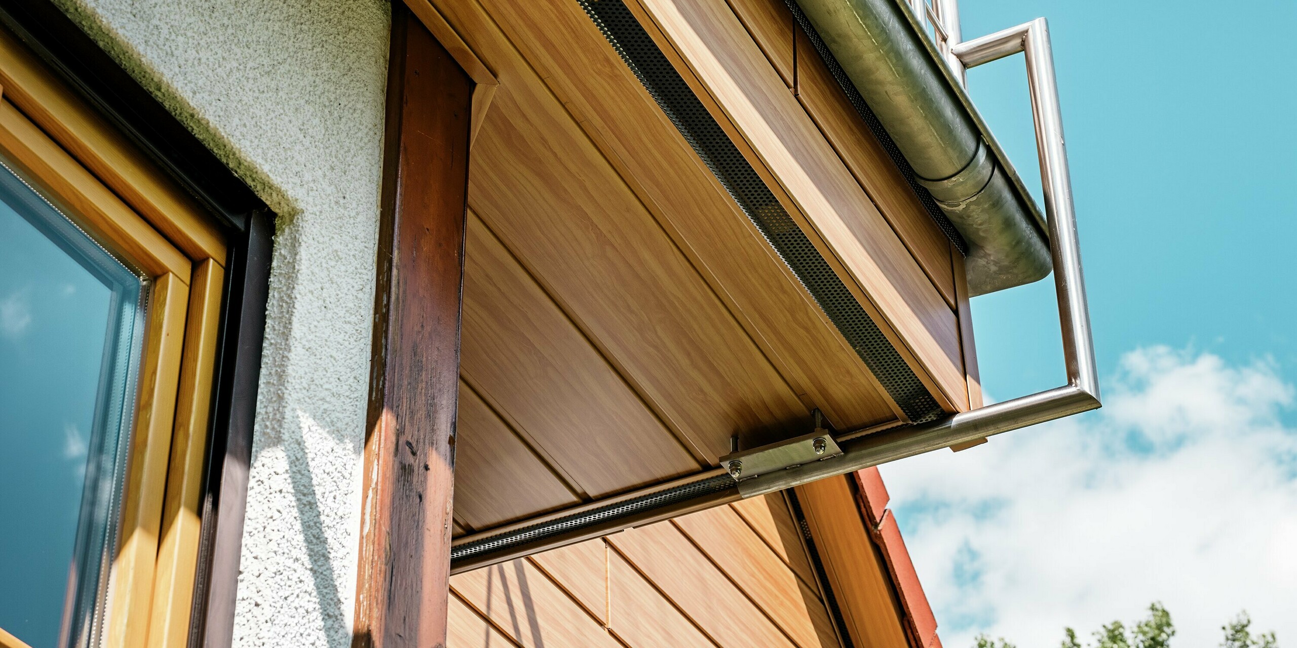 L'image présente un gros plan dynamique du pignon rénové d'une maison à Blieskastel, revêtu de Siding PREFA en chêne naturel. Vue de dessous, la façade révèle des panneaux imitation bois d'une authenticité et d'une qualité exceptionnelles, tandis que la délicate rambarde métallique du balcon semble flotter avec élégance. Les tons chauds de la façade en aluminium s'harmonisent avec le ciel ensoleillé et la végétation luxuriante au premier plan. Grâce à la surface PREFA, résistante aux intempéries et indéformable, l'aspect naturel est préservé durablement – ​​un détail raffiné qui met en valeur l'ensemble du bâtiment.