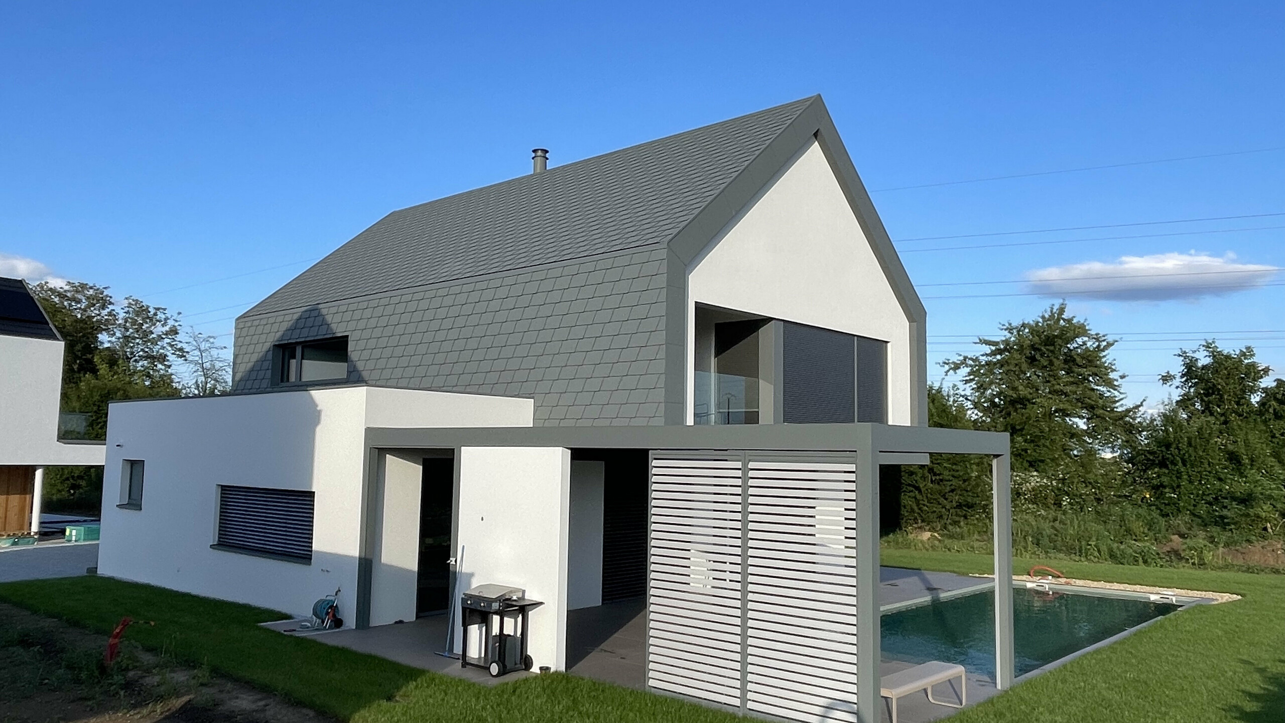 Vue d'ensemble de cette nouvelle maison individuelle dont la toiture et la façade contemporaine ont été recouvertes de bardeaux en aluminium PREFA. On aperçoit une pergola moderne à côté d'une piscine en premier plan.