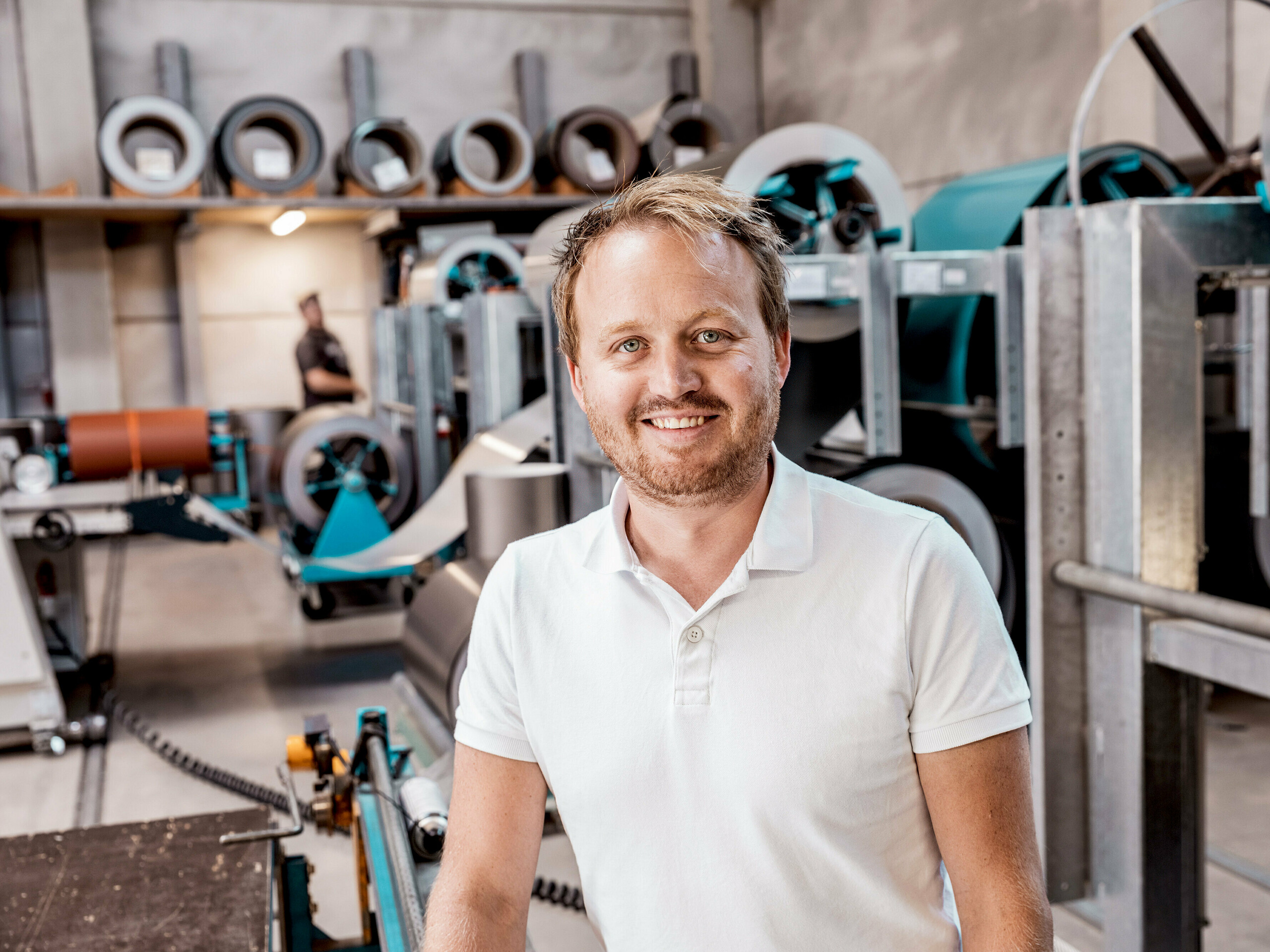 La photo montre David Thumfart, tôlier chez Kapl Bau GmbH, entreprise spécialisée dans la toiture et la tôlerie, dans son atelier devant les machines de traitement des bobines de métal. Souriant et professionnel, il se tient fièrement au milieu des équipements de production ultramodernes utilisés pour la fabrication de systèmes de façade et de toiture en aluminium. Les machines en arrière-plan témoignent de la précision requise pour fabriquer des produits de haute qualité comme la façade PREFALZ, utilisée sur une maison individuelle à Freistadt. David Thumfart incarne le dévouement et le savoir-faire nécessaires à la production et à l'installation d'éléments de construction durables et esthétiques.