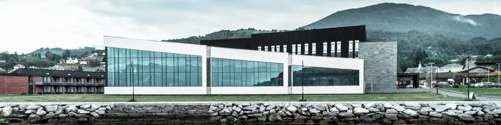 Dans ce cliché, nous voyons les reflets de la façade du centre de santé Hardangerbadet à Oystese dans l'eau. Celle-ci a été recouverte en losange de façade 44x44 en aluminium naturel. On peut apercevoir un bout du Fjord Hardangerfjord dans le fond.