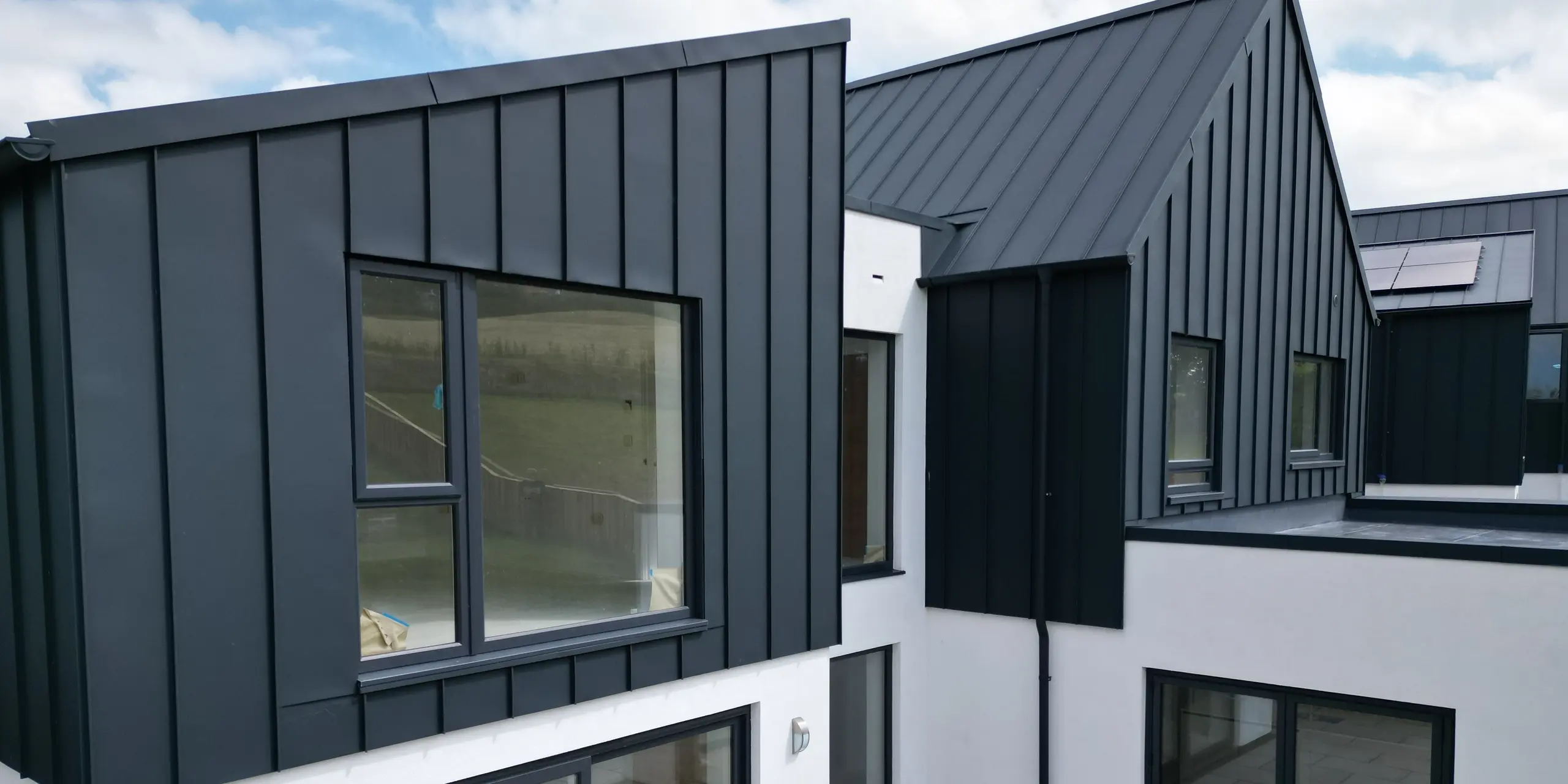 Vue détaillée d'une maison individuelle avec jardin et terrasse, recouverte de PREFALZ en P.10 anthracite. La toiture à joint debout en aluminium allie design moderne et durabilité et souligne l'approche de construction durable. Les grandes fenêtres apportent beaucoup de lumière à l'intérieur.