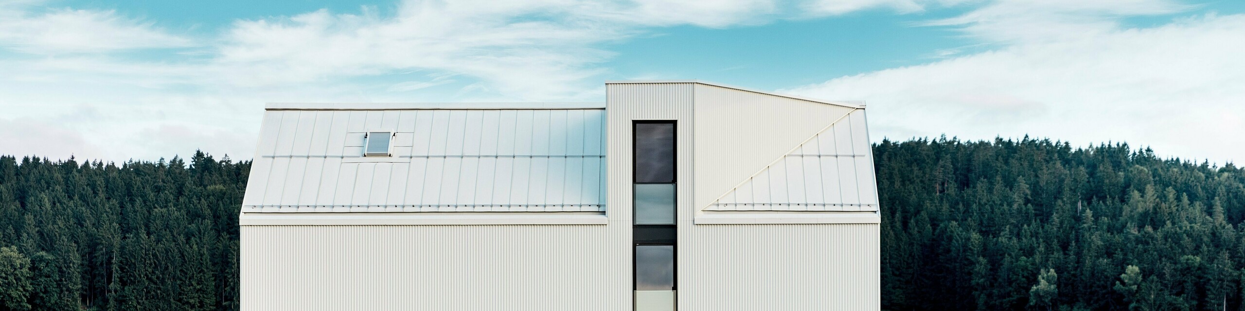 L'image présente une maison individuelle à Freistadt, vue de profil, mettant en valeur son architecture minimaliste et remarquable. La façade en aluminium blanc pur PREFALZ et le toit à pignon abrupt dessinent une silhouette épurée et moderne qui s'intègre harmonieusement à la verdure environnante. Les grandes fenêtres et l'élégante lucarne soulignent la fonctionnalité du design, tandis que la qualité de l'aluminium garantit à la fois une esthétique durable et une excellente résistance aux intempéries. Par sa forme simple et l'utilisation de matériaux pérennes, le bâtiment dégage une élégance intemporelle et représente une parfaite harmonie entre architecture et nature.