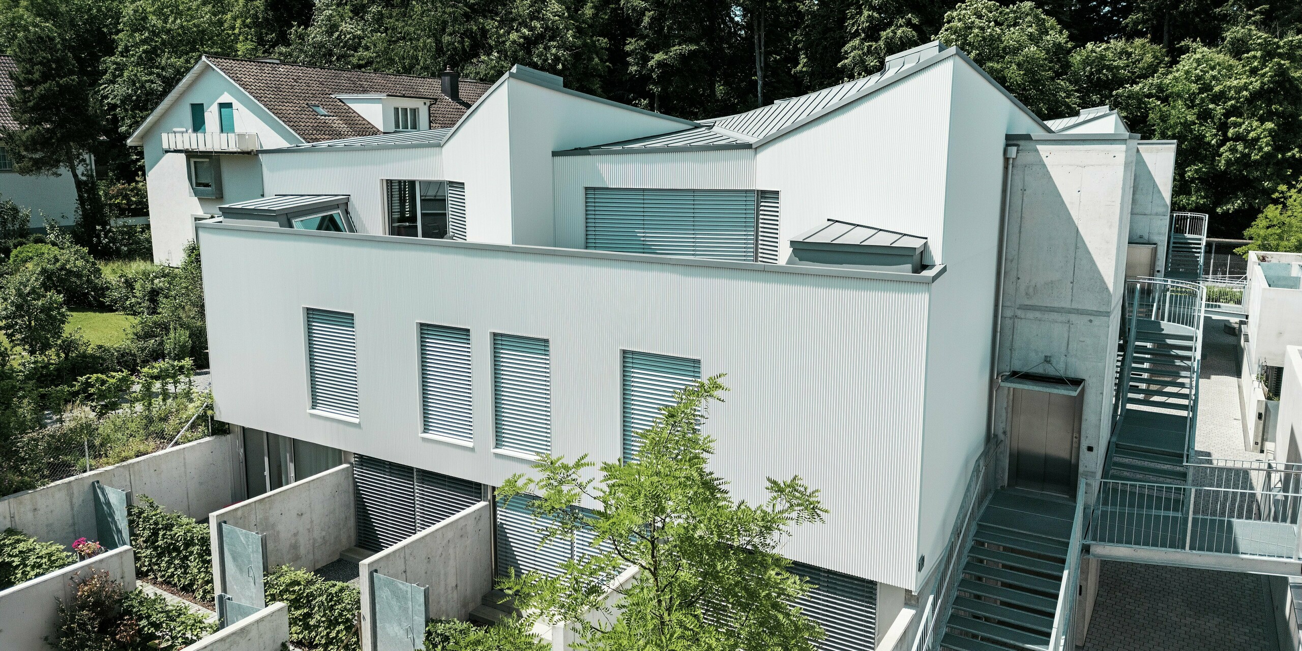 Vue aérienne d'un ensemble de mini-maisons modernes à Zollikerberg, caractérisées par des façades à profil dentelé PREFA P.10 blanc pur et des toitures PREFALZ gris quartz. La pureté des lignes et le contraste des matériaux confèrent au projet une esthétique remarquable. L'aluminium de haute qualité garantit durabilité, protection contre les intempéries et précision architecturale dans un espace réduit.