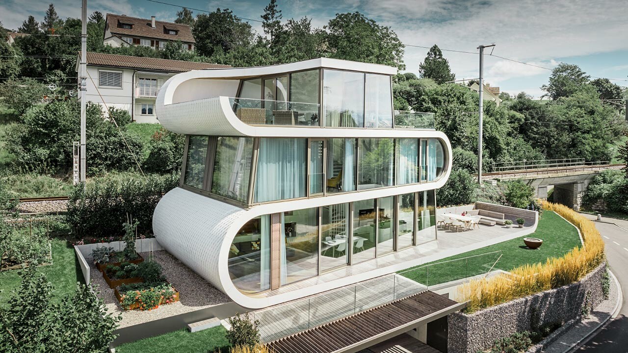 Photo de la Flexhouse moderne au bord du lac de Zurich. Avec sa forme hors du commun, la Flexhouse attire toujours le regard. Les séparations entre étages s’étirent comme une bande blanche ondulée à travers le bâtiment. Les éléments de façade arrondis ont été recouverts de bardeaux PREFA en aluminium blanc pur.