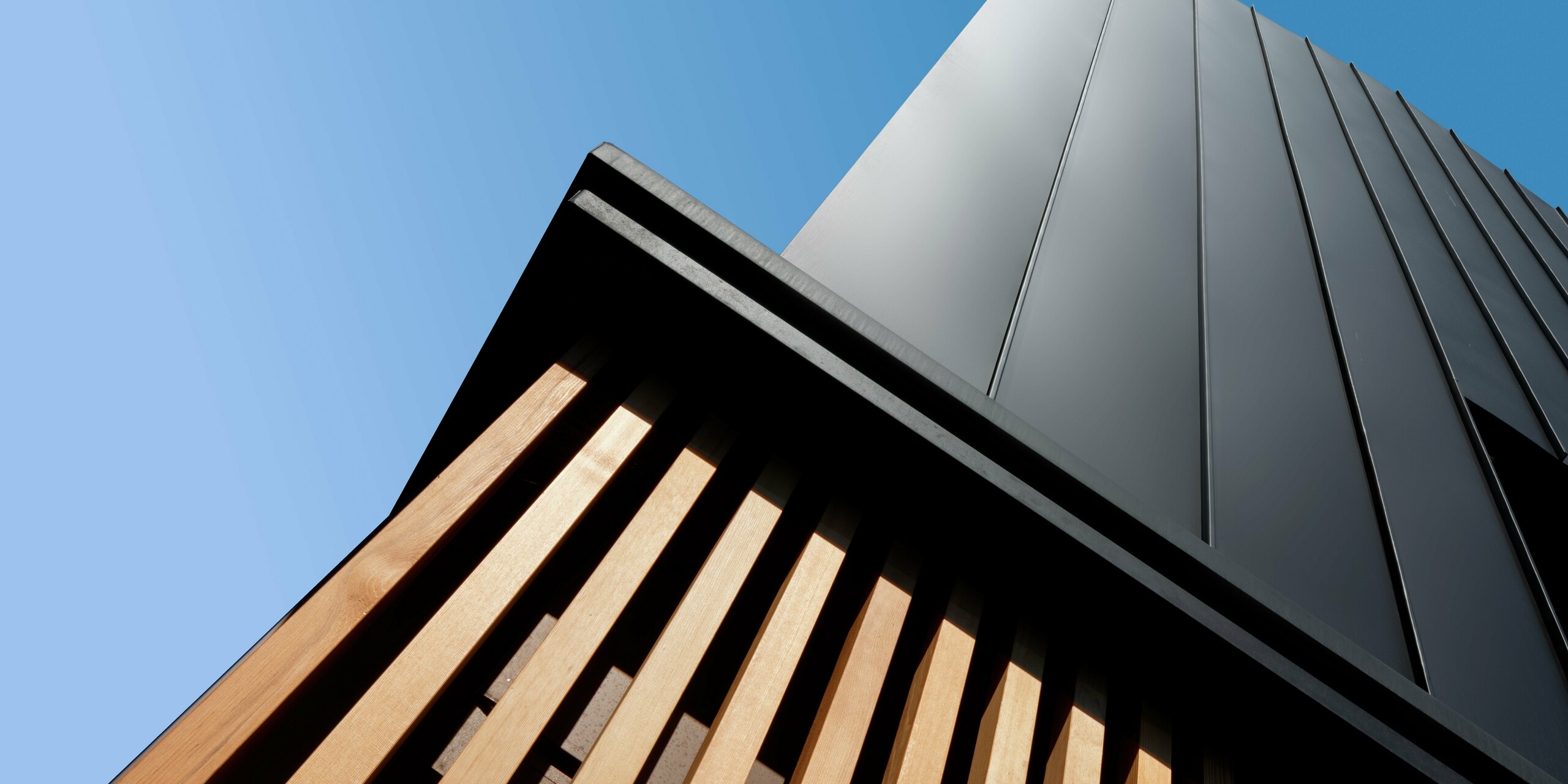 Détail architectural d'une maison jumelée moderne à Cattolica : un bardage vertical en aluminium PREFALZ gris noir s'harmonise avec d'élégantes lames de bois. La pureté des lignes et la qualité de la surface PREFA garantissent durabilité, protection contre la corrosion et un design saisissant sous un ciel d'azur.