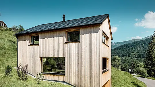La lumière du soleil dessine des ombres marquées sur la façade verticale en sapin blanc de la maison Immi, qui s’intègre harmonieusement au versant escarpé d’Ebnit. Au-dessus de cette architecture en bois aux lignes épurées se déploie le toit solaire PREFA noir, composé de tuiles solaires intégrées et de panneaux de toiture R.16. Cette solution robuste en aluminium produit de l’électricité, assure une protection durable et préserve visuellement la silhouette épurée du bâtiment.