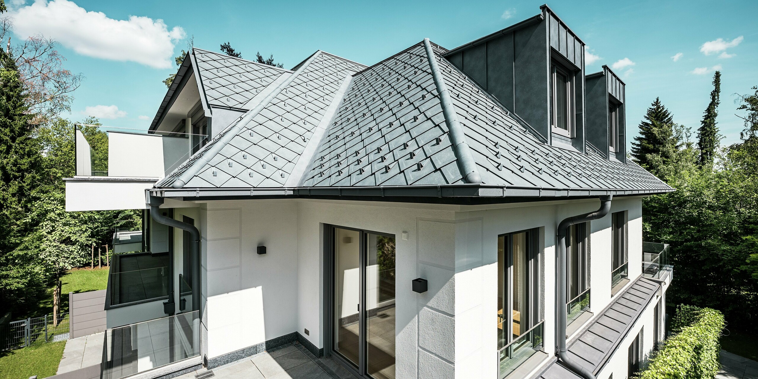 Maison multifamiliale moderne à l'architecture de toiture remarquable, réalisée en PREFA P.10 Gris Pierre : la surface du toit est recouverte de losanges PREFA 29 x 29 cm de haute qualité, créant un aspect texturé et élégant. Des lucarnes plates stylées en PREFALZ s'intègrent harmonieusement au design. Des arrêts de neige intégrés et un système d'évacuation des eaux pluviales PREFA esthétique, avec gouttières et descentes, soulignent la sophistication technique. La toiture en aluminium durable résiste à toutes les conditions climatiques et allie esthétique et fonctionnalité – idéale pour la construction résidentielle contemporaine.