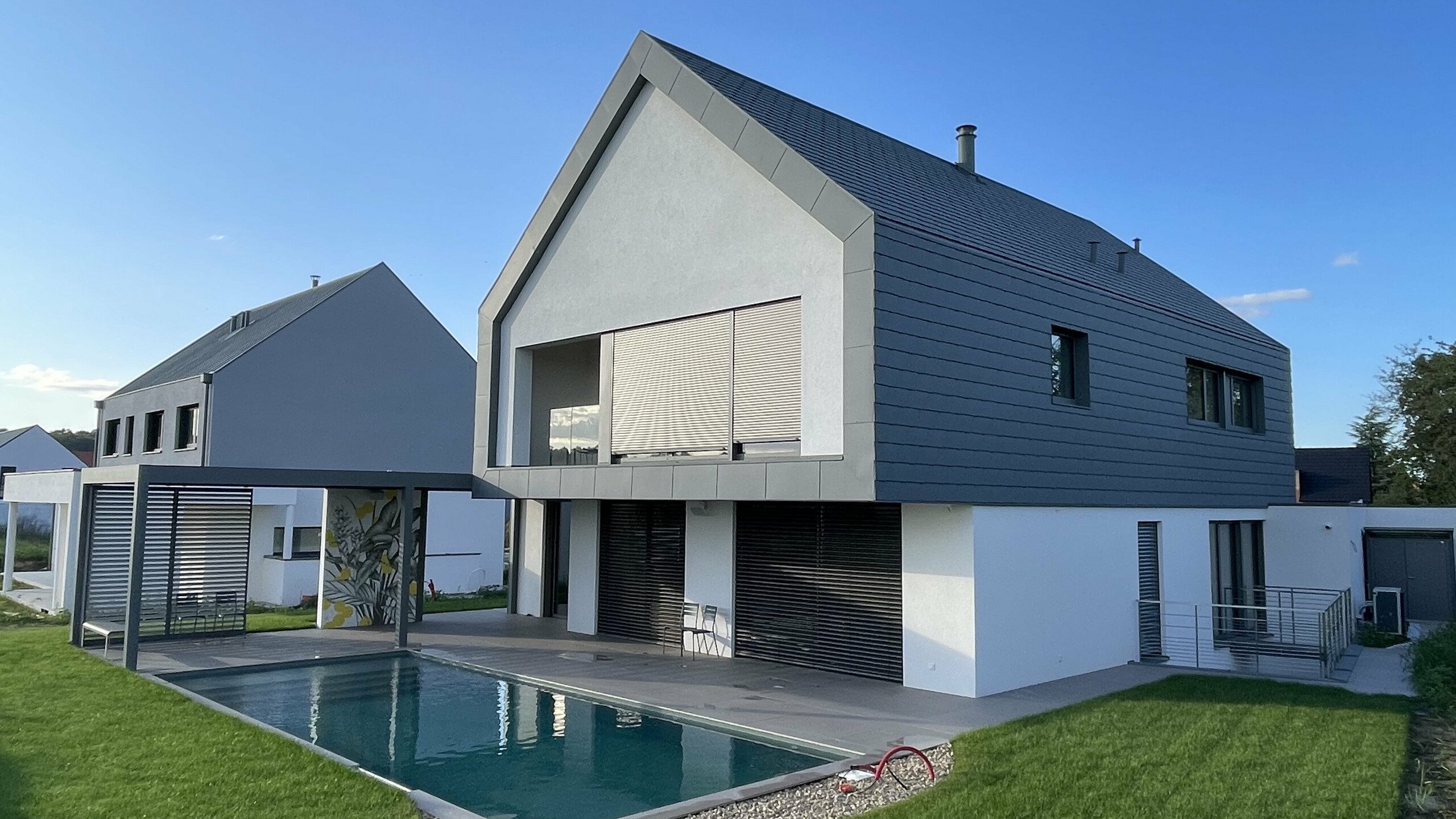 Vue d'ensemble de cette nouvelle maison individuelle dont la toiture et la façade contemporaine ont été recouvertes de bardeaux en aluminium PREFA. On aperçoit une jolie piscine en premier plan.