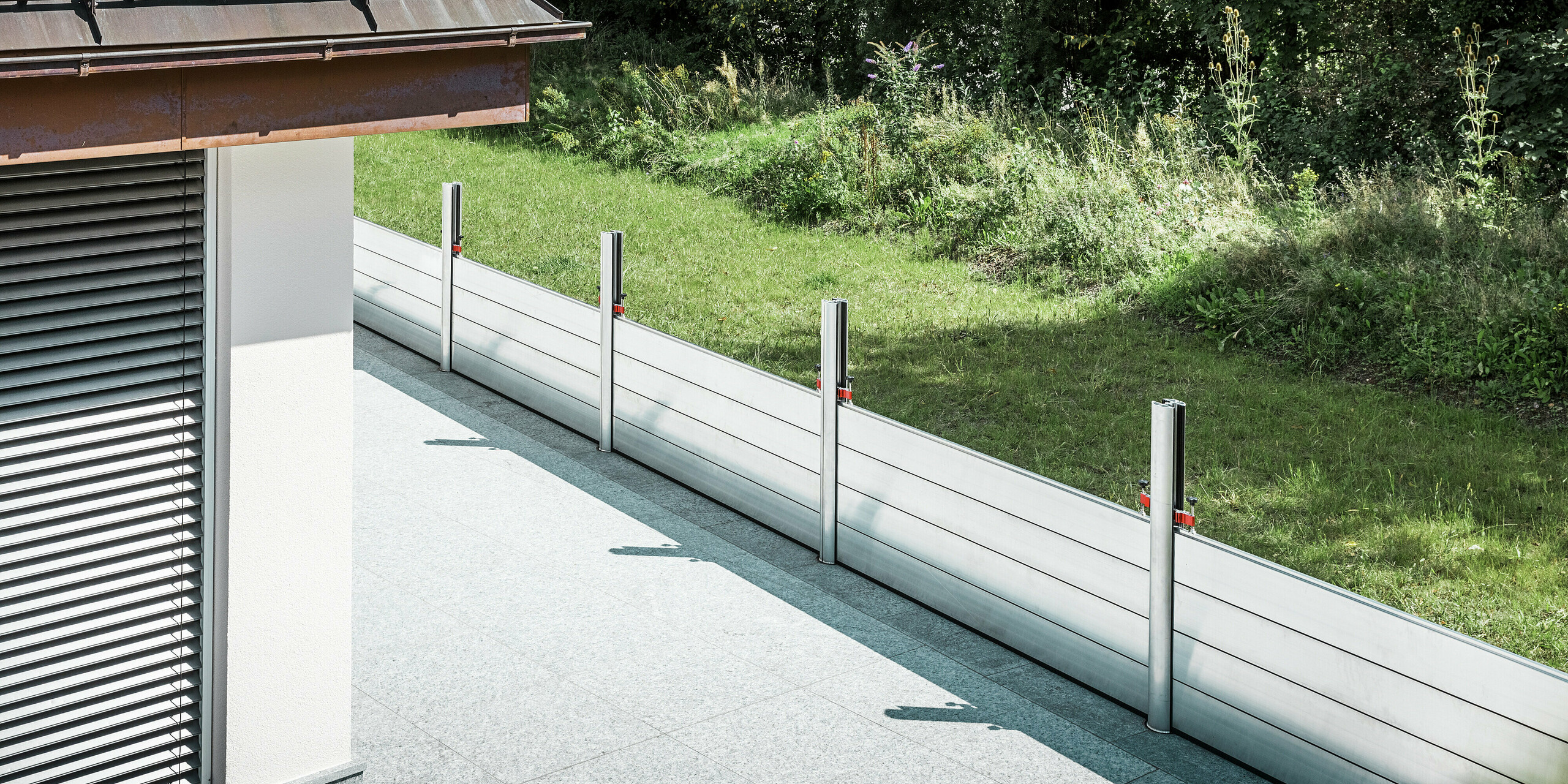 Vue oblique du système de protection contre les crues PREFA installé le long de la terrasse d'un immeuble résidentiel au Tyrol. Les batardeaux robustes en aluminium sont montés entre des profilés de support ancrés à demeure, eux-mêmes solidement ancrés au sol par des ancrages au sol. Ce système mobile de protection contre les crues offre une protection fiable contre les inondations potentielles provenant de l'espace vert adjacent. La façade articulée avec stores est visible au premier plan, tandis que le système de protection s'intègre discrètement et fonctionnellement dans l'environnement résidentiel de qualité.