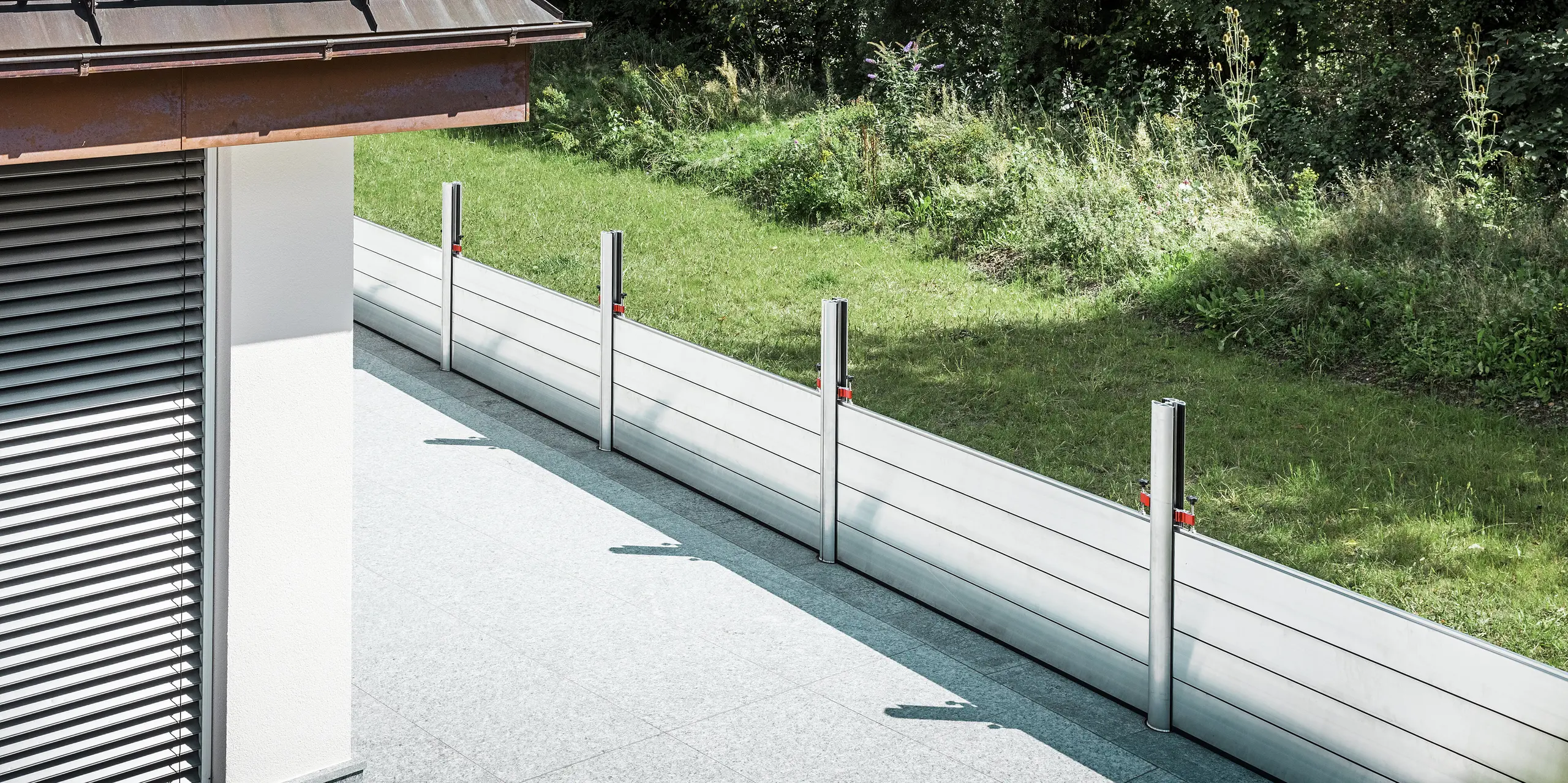 Vue oblique du système de protection contre les crues PREFA installé le long de la terrasse d'un immeuble résidentiel au Tyrol. Les batardeaux robustes en aluminium sont montés entre des profilés de support ancrés à demeure, eux-mêmes solidement ancrés au sol par des ancrages au sol. Ce système mobile de protection contre les crues offre une protection fiable contre les inondations potentielles provenant de l'espace vert adjacent. La façade articulée avec stores est visible au premier plan, tandis que le système de protection s'intègre discrètement et fonctionnellement dans l'environnement résidentiel de qualité.