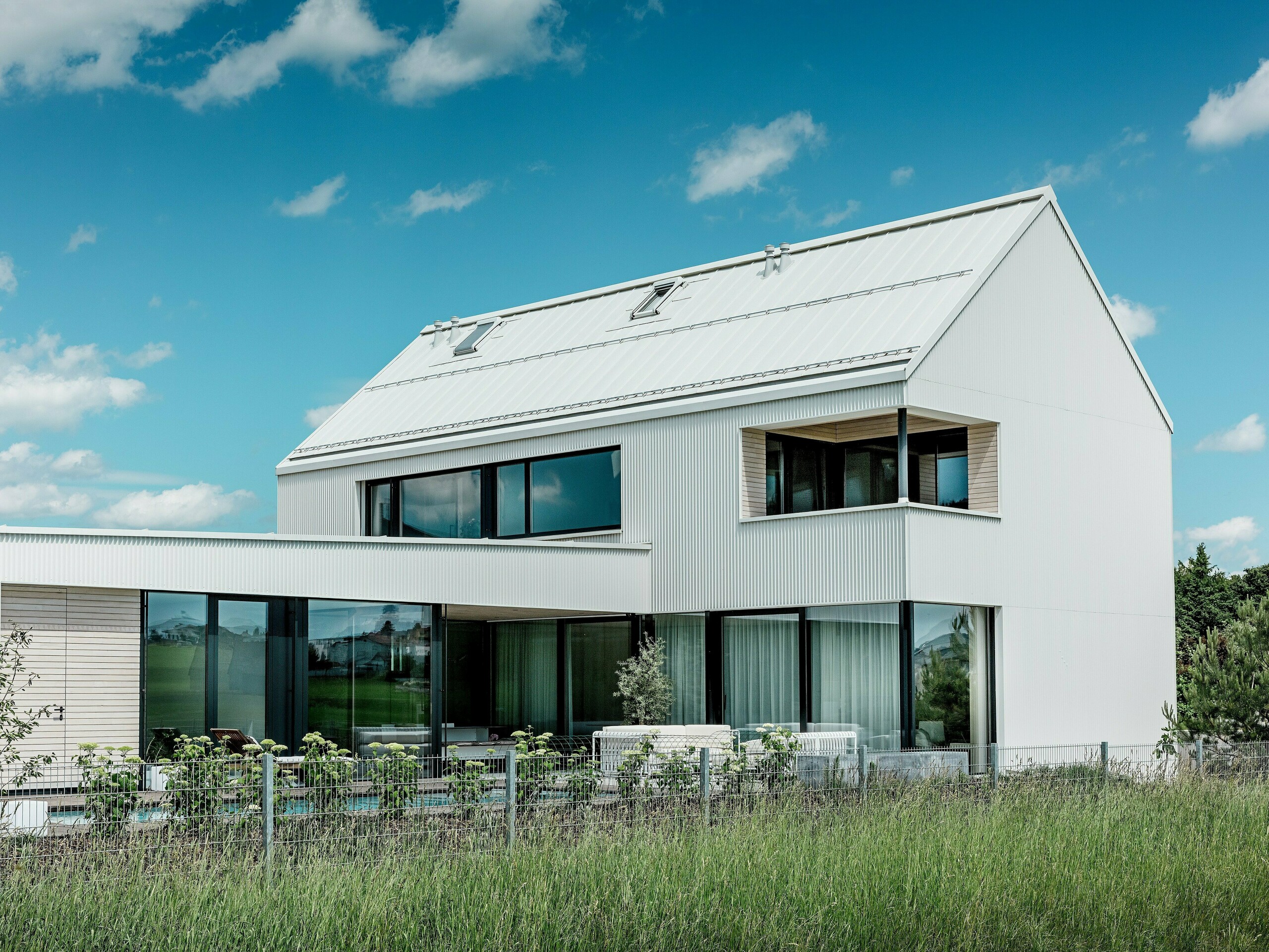 Cette impressionnante maison individuelle à Freistadt séduit par son architecture élégante, sublimée par la façade et la toiture en aluminium PREFALZ d'un blanc pur. La façade en zigzag crée une silhouette géométrique et moderne qui s'intègre harmonieusement au paysage environnant. De larges baies vitrées inondent l'intérieur de lumière naturelle et offrent de vastes vues sur le paysage. Le toit à forte pente et le balcon moderne habillé de bois soulignent le design minimaliste et la construction durable de la maison. PREFALZ garantit une durabilité visuelle et une résistance aux intempéries optimales, assurant une longue durée de vie et un entretien minimal.