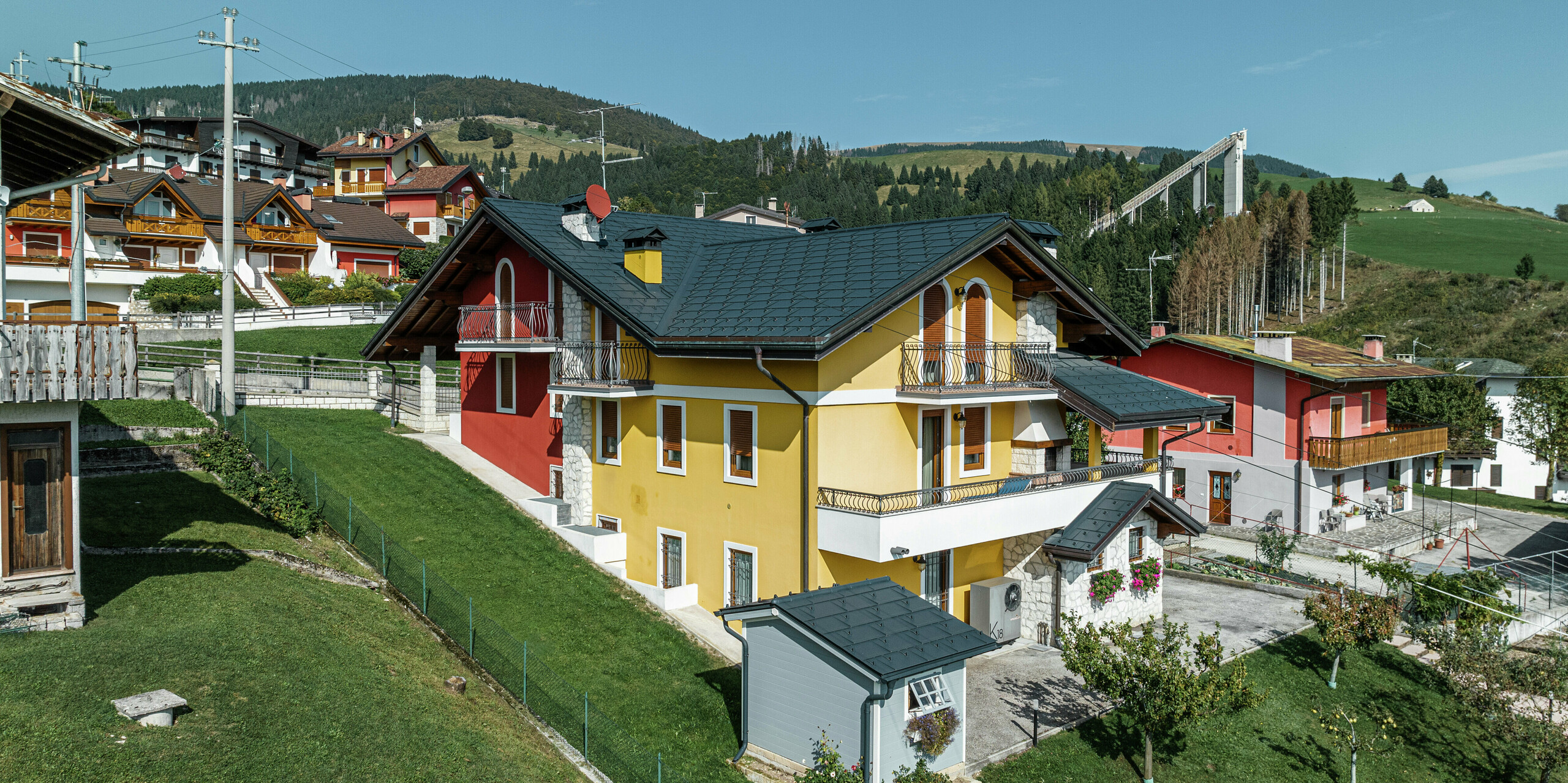 Maison individuelle à Gallio dans une architecture alpine traditionnelle avec une façade en plâtre jaune-rouge vif et des éléments en pierre naturelle. Le R.16 PREFA en P.10 gris sombre offre une résistance maximale aux intempéries et s'harmonise parfaitement avec l'environnement. Le système de gouttière avec gouttière suspendue et tuyau de descente PREFA – également en gris sombre P.10 – assure une protection fiable. Entourée de prairies verdoyantes et surplombant les montagnes, la maison allie esthétique, durabilité et fonctionnalité. En arrière-plan, vous pouvez voir le célèbre tremplin de saut à ski de la région.