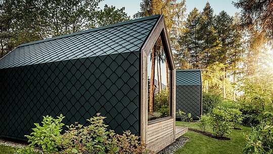 Une minuscule maison minimaliste, nichée dans la verdure du Secret Koi Garden sur les rives du lac Natterer, se pare d'une douce lumière du soir. Toit et façade sont entièrement revêtus de losanges PREFA en aluminium P.10 anthracite, dont la texture précise, évoquant des écailles de poisson, crée des accents saisissants. Durable et résistant aux intempéries, l'aluminium PREFA protège efficacement la structure et s'harmonise parfaitement avec le bois, le verre et la nature environnante.