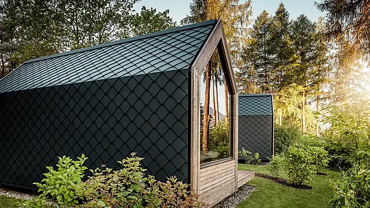 Une minuscule maison minimaliste, nichée dans la verdure du Secret Koi Garden sur les rives du lac Natterer, se pare d'une douce lumière du soir. Toit et façade sont entièrement revêtus de losanges PREFA en aluminium P.10 anthracite, dont la texture précise, évoquant des écailles de poisson, crée des accents saisissants. Durable et résistant aux intempéries, l'aluminium PREFA protège efficacement la structure et s'harmonise parfaitement avec le bois, le verre et la nature environnante.