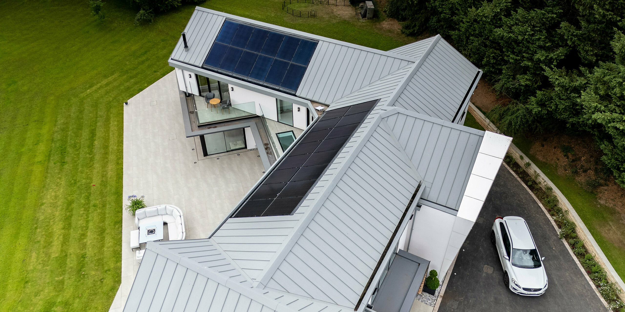 Cette villa moderne de Draycott-in-the-Clay est sublimée par une élégante toiture PREFALZ gris quartz et un système photovoltaïque côté jardin. Sa structure à joints debout épurée lui confère un design intemporel et futuriste qui s'intègre harmonieusement à la végétation environnante. La toiture métallique haut de gamme est non seulement esthétique, mais contribue également à l'efficacité énergétique de la propriété. L'alliance de matériaux nobles comme le métal et la technologie solaire fait de cette maison une pionnière en matière de construction durable et élégante. Une voiture blanche est garée devant le bâtiment, comme pour accueillir des visiteurs.