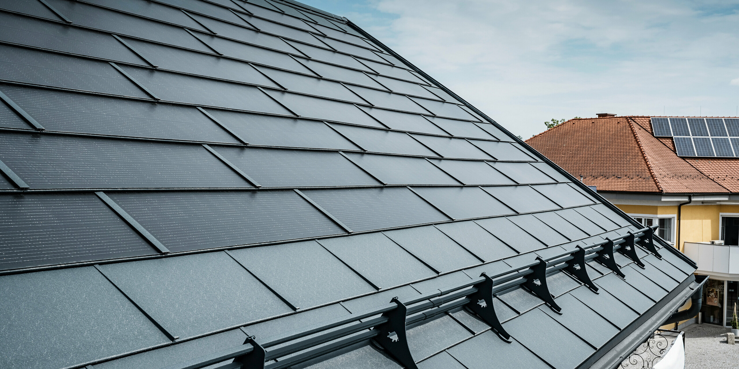 Vue d'une toiture moderne avec des tuiles solaires PREFA de couleur anthracite. Près de l'avant-toit, le toit a été équipé de R.16 classiques et d'un système de protection contre la neige PREFA. En arrière-plan, le toit d'une maison voisine est visible avec des tuiles traditionnelles et des panneaux solaires sur le toit, soulignant le contraste entre la conception d'un nouveau toit et d'un toit plus traditionnel. Le tuile solaire innovante de PREFA allie esthétique et fonctionnalité et offre une solution énergétique durable pour tous les types de bâtiments.