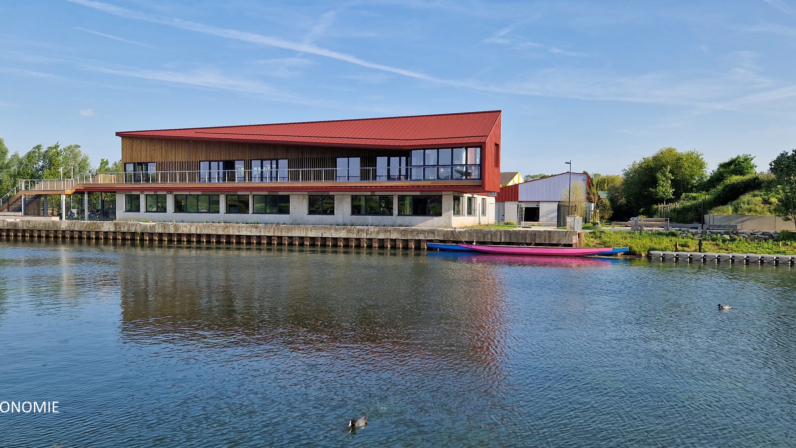 Vue éloignée du stade nautique d'Arras, la teinte rouge oxyde de l'aluminium Prefa se reflète dans la rivière