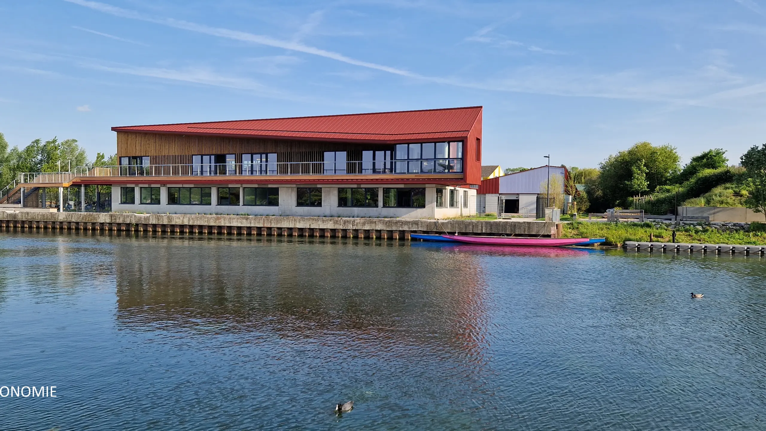 Vue éloignée du stade nautique d'Arras, la teinte rouge oxyde de l'aluminium Prefa se reflète dans la rivière