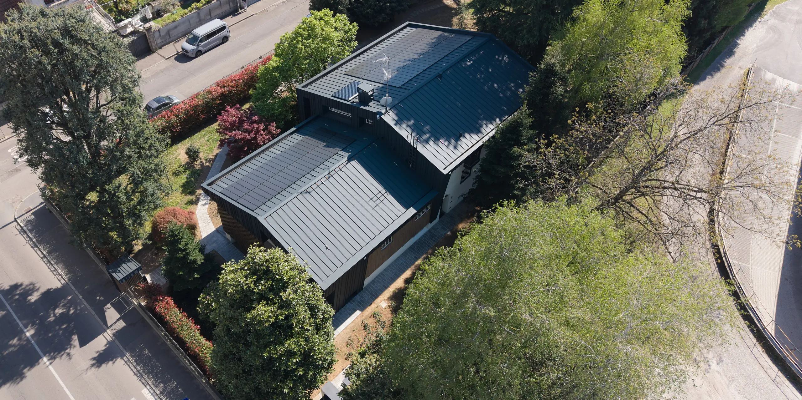 La photographie aérienne révèle la Villa Castellanza, une demeure moderne dotée d'une remarquable enveloppe en aluminium PREFA. La toiture PREFA aux lignes élégantes et la façade PREFA anthracite P.10, parfaitement intégrée, attirent particulièrement le regard et confèrent à la résidence un style architectural épuré et contemporain. Les modules solaires PREFA, largement intégrés, se fondent harmonieusement dans le paysage des deux toitures à deux pans et soulignent l'engagement de la villa en faveur d'une énergie durable. Entourée d'arbres matures, la villa offre un contraste saisissant entre nature et architecture moderne. Durables et nécessitant peu d'entretien, les solutions en aluminium PREFA allient design sophistiqué, fonctionnalité et efficacité énergétique optimale.