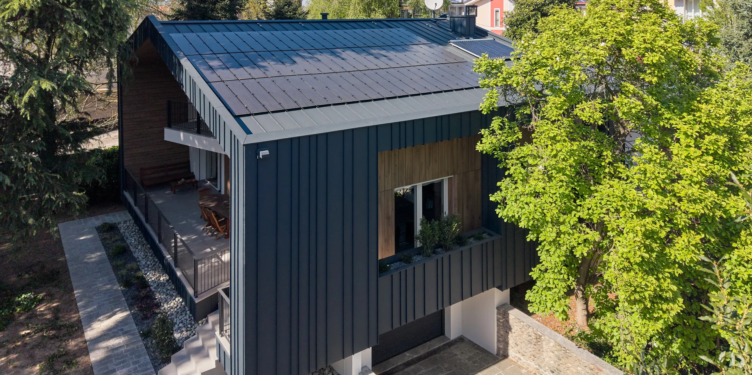 Vue détaillée de la Villa Castellanza, avec sa toiture et sa façade continues en PREFALZ couleur Anthracite P.10. De grands modules solaires PREFALZ intégrés définissent l'enveloppe moderne du bâtiment et lui confèrent une esthétique harmonieuse. Les produits en aluminium PREFALZ, d'une grande durabilité, allient élégance architecturale, production d'énergie durable et haute résistance. Entourée d'arbres, la maison est particulièrement impressionnante.