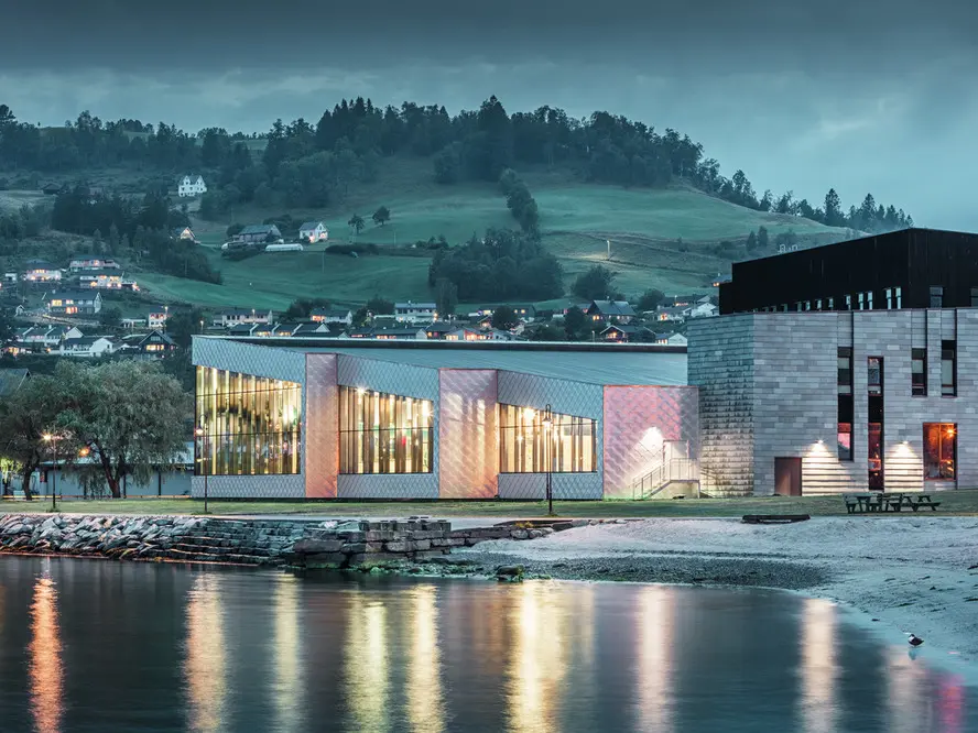Dans ce cliché, nous voyons les reflets de la façade du centre de santé Hardangerbadet à Oystese dans l'eau. Celle-ci a été recouverte en losange de façade 44x44 en aluminium naturel. On peut apercevoir un bout du Fjord Hardangerfjord dans le fond.