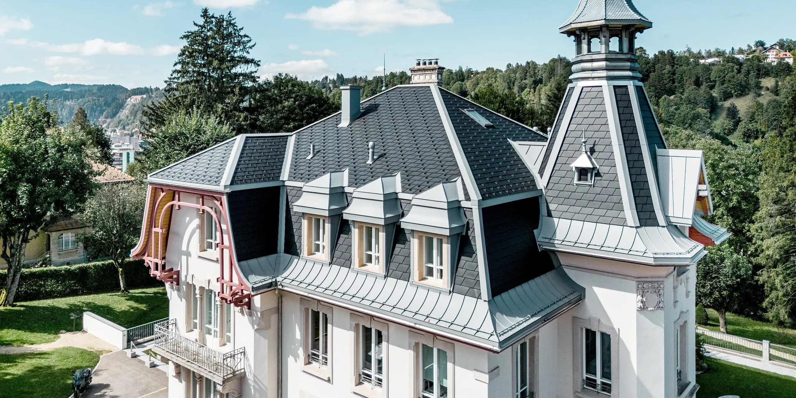Dynamische Perspektive auf die Manufacture Horlogère du Locle in der Schweiz mit markantem Turm und kunstvoll gegliederter Dachlandschaft. Die steilen, facettenreichen Flächen mit PREFA Dachschindeln in P.10 Schwarz setzen starke geometrische Akzente, während PREFALZ in Patinagrau elegante Schwünge und präzise Anschlüsse an Gauben und Rundungen ermöglicht. Das leichte Aluminiumdach überzeugt durch hohe Langlebigkeit, Witterungsbeständigkeit und maximale Flexibilität – eine perfekte Verbindung aus ästhetischer Architektur, technischer Präzision und nachhaltigem Werterhalt.