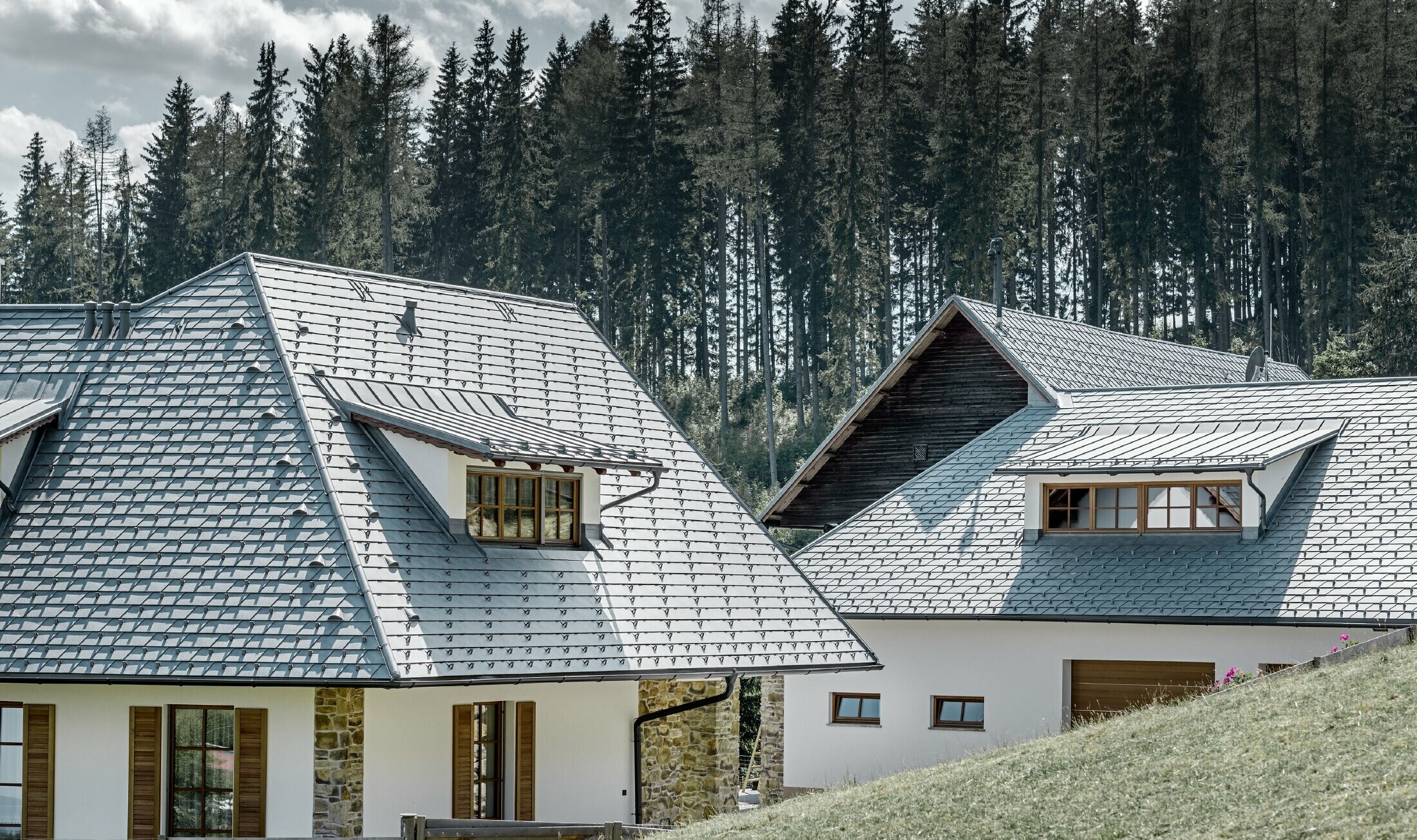 Vue de derrière de la maison individuelle construite à flanc de coteau ; la toiture a été recouverte de bardeaux de toiture en alu couleur gris pierre, les lucarnes rampantes avec couverture à joints debout, également en gris pierre. On peut voir une forêt en arrière plan.