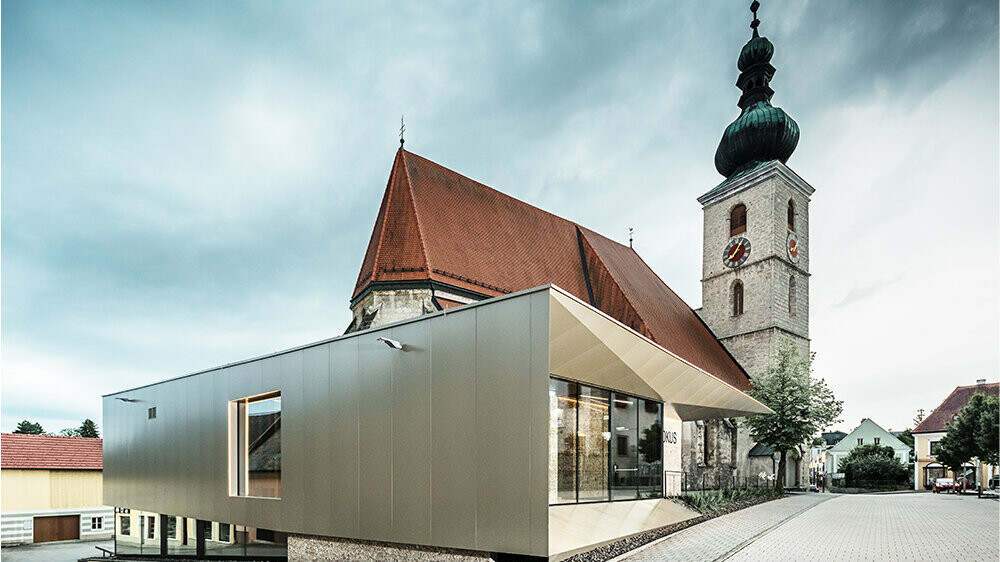 Das moderne Pfarrzentrum in Sierning wurde an Fassade und Untersicht mit PREFABOND Aluminium Verbundplatten in Bronze verkleidet. Im Hintergrund hebt sich die historische Kirche ab - ein schöner Kontrast zwischen Geschichte und Moderne.