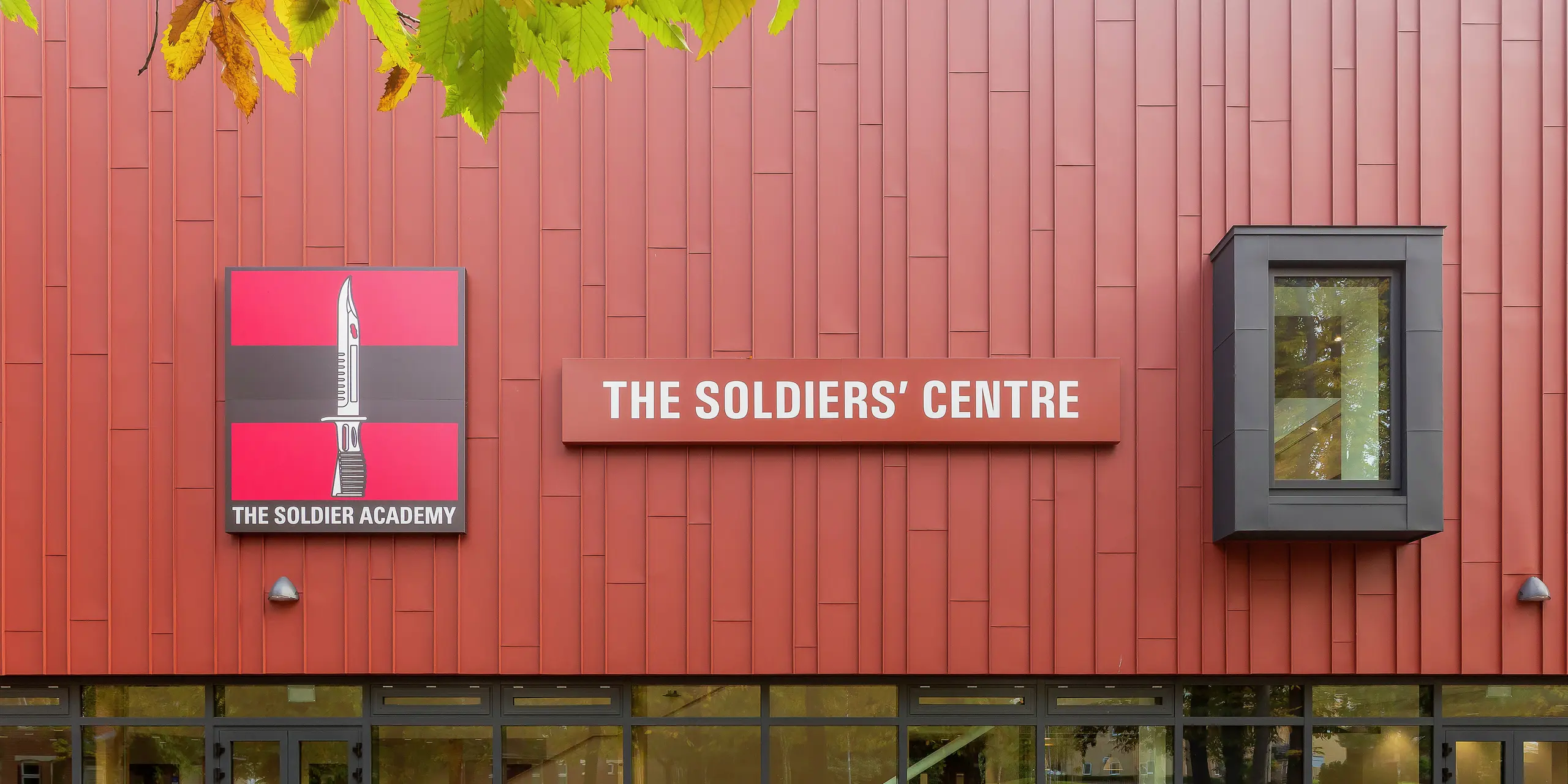 Bâtiment social construit spécialement pour l'armée avec façade PREFALZ : La toiture à joint debout traitée avec précision en P.10 rouge oxyde confère au bâtiment une apparence remarquable. La zone d'entrée impressionne par ses généreuses façades vitrées qui offrent luminosité et transparence. Le logo « Soldier Academy » et le panneau de bâtiment accrocheur créent des accents visuels clairs. Les cadres de fenêtre en P.10 anthracite complètent harmonieusement le design. Produits en aluminium PREFA de haute qualité pour une architecture durable et esthétique.